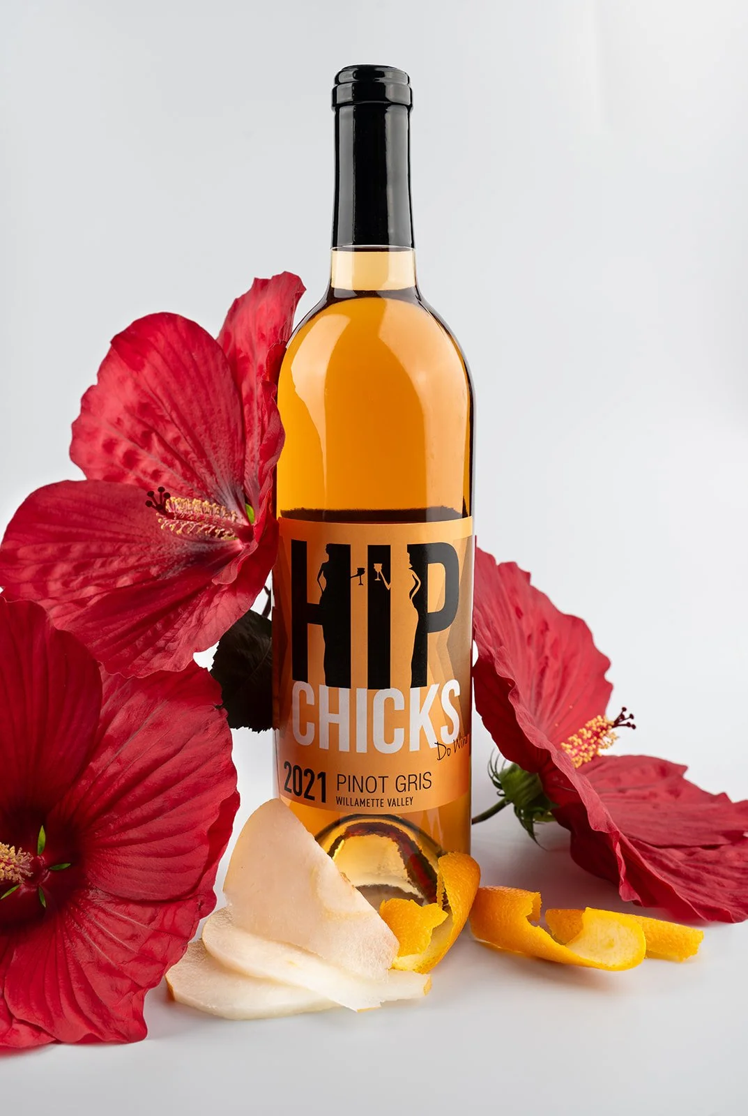 A bottle of wine labeled "Hips Chicks" 2021 Pinot Gris from Willamette Valley, surrounded by red hibiscus flowers, a white pear, and lemon peel against a plain background.