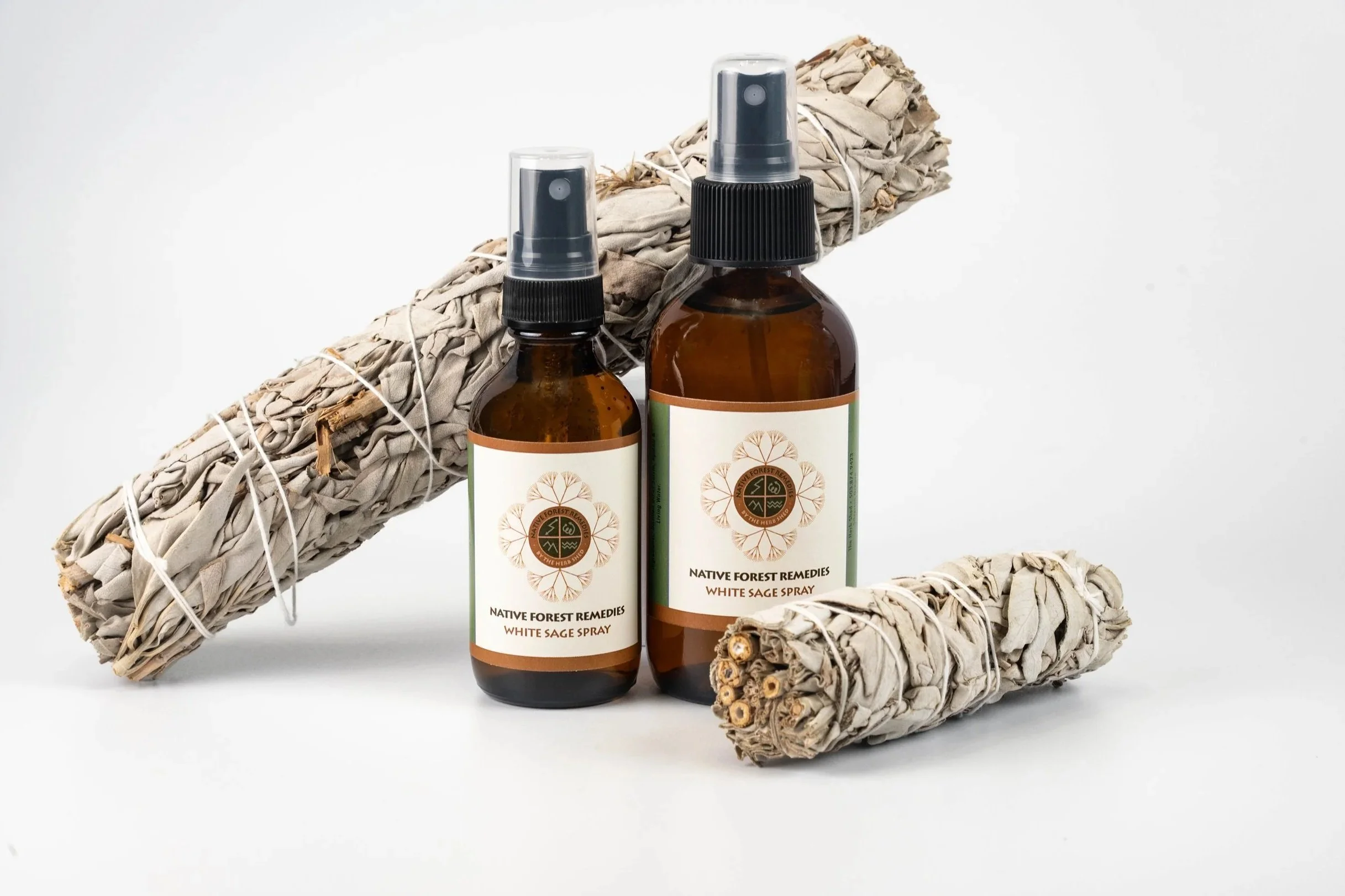 Two amber glass spray bottles labeled 'Native Forest Remedies White Sage Spray,' placed with two bundles of white sage smudge sticks wrapped in string on a white background.