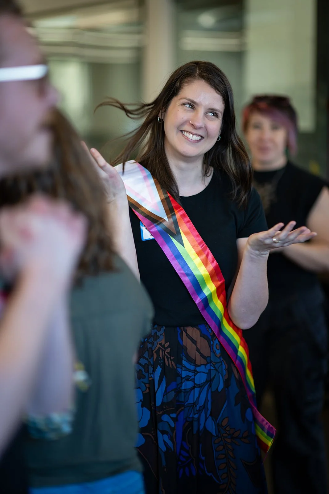 A woman with shoulder-length dark hair, wearing a black shirt and a rainbow sash, is smiling and gesturing with her hand, during a social gathering. There is another person blurred in the foreground on the left and a person with pink hair in the background.