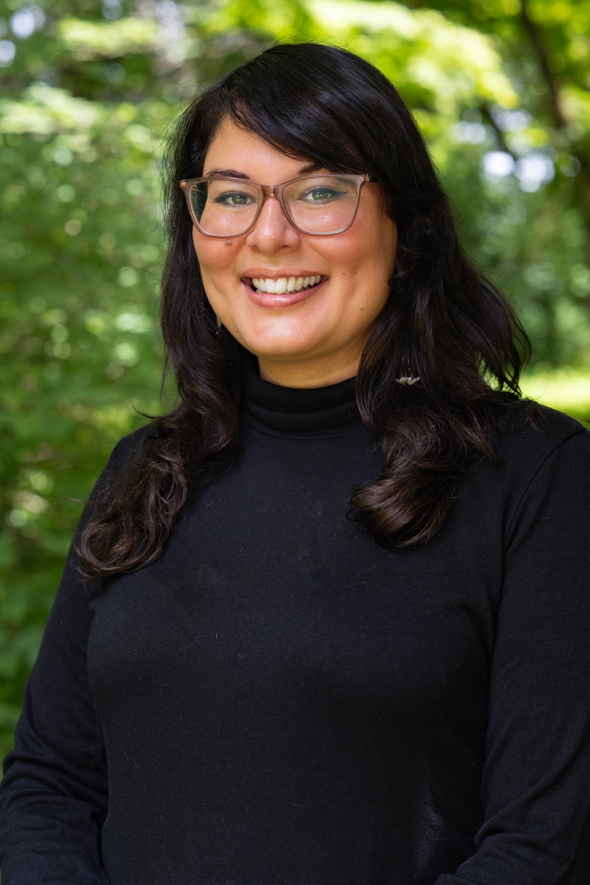 A woman with glasses, long dark hair, and a black turtleneck smiling outdoors with a background of green trees.