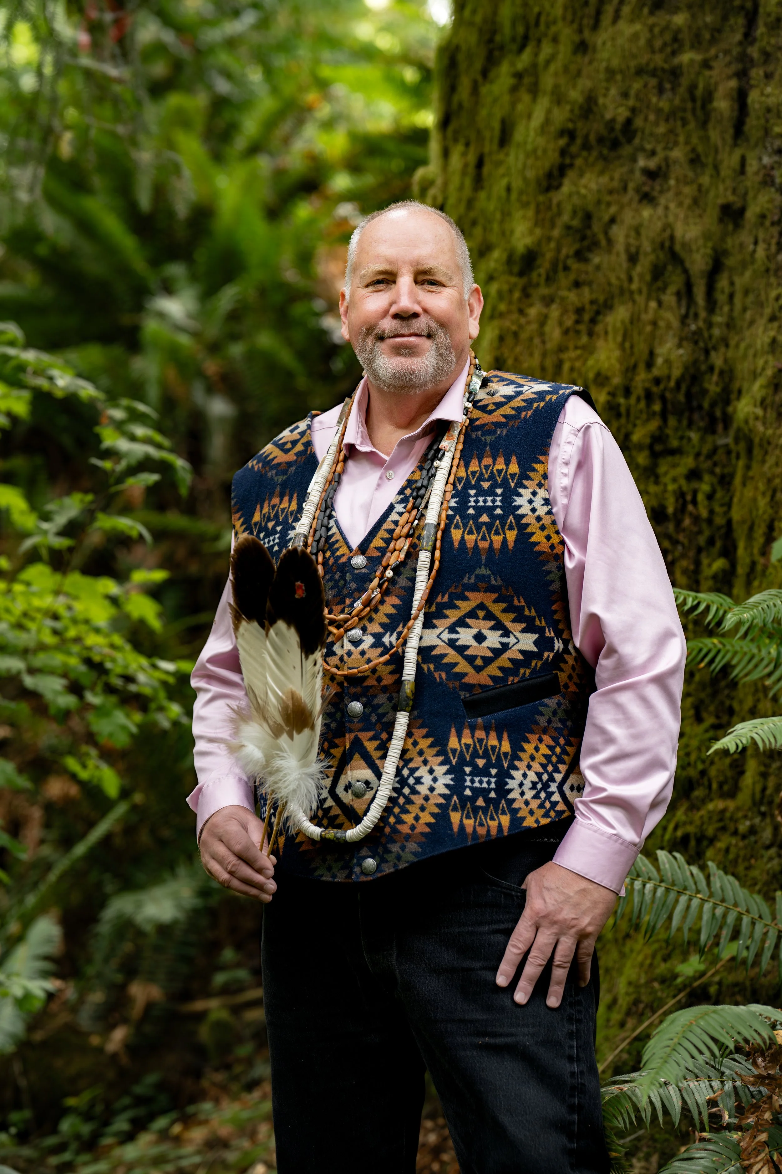 A man standing outdoors in a forest, wearing a traditional patterned vest over a pink shirt, holding a feather fan, and adorned with beaded necklaces.