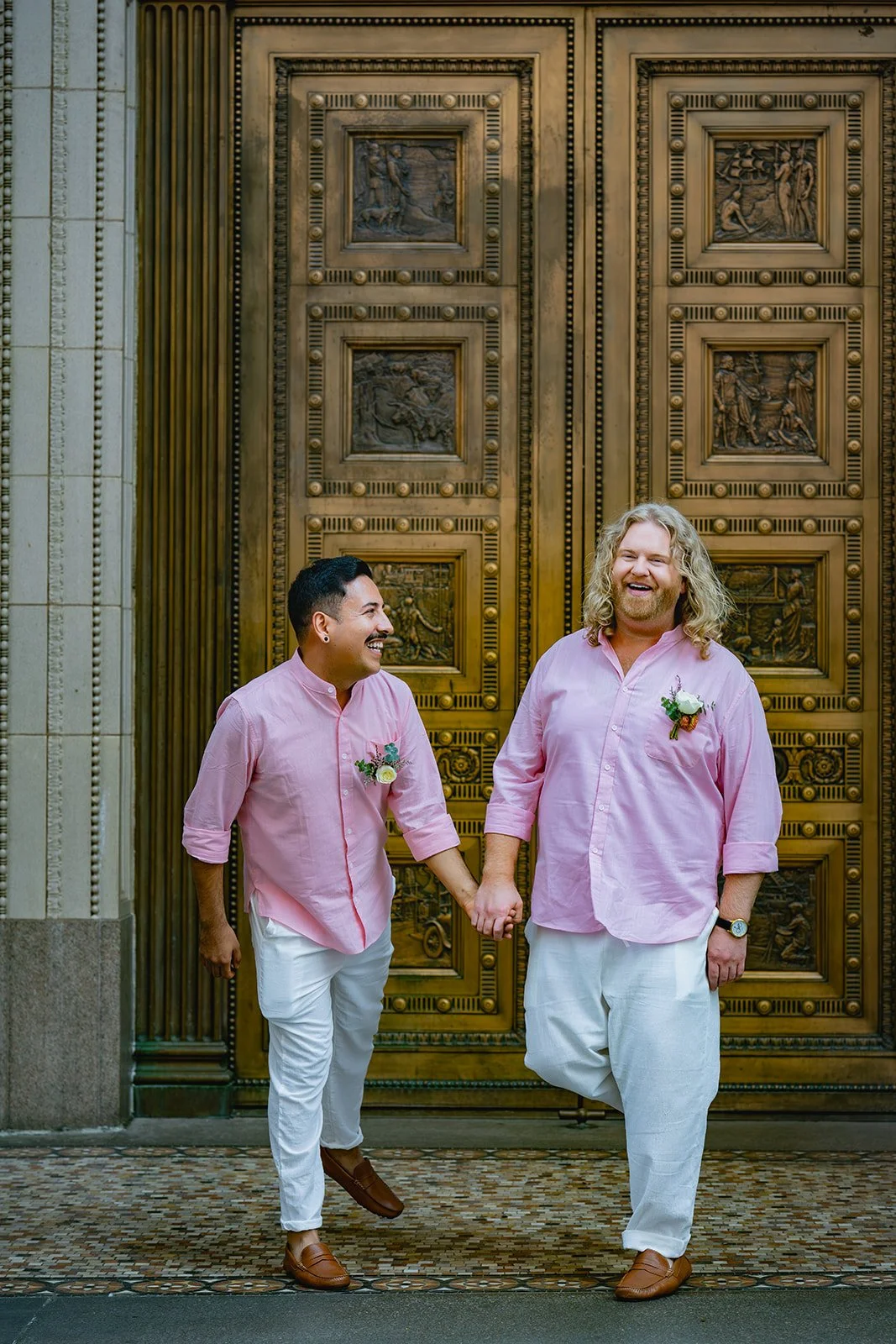 A same-sex couple holding hands and smiling in front of an ornate, gold-colored door, dressed in pink shirts and white pants, with boutonnieres pinned to their shirts.