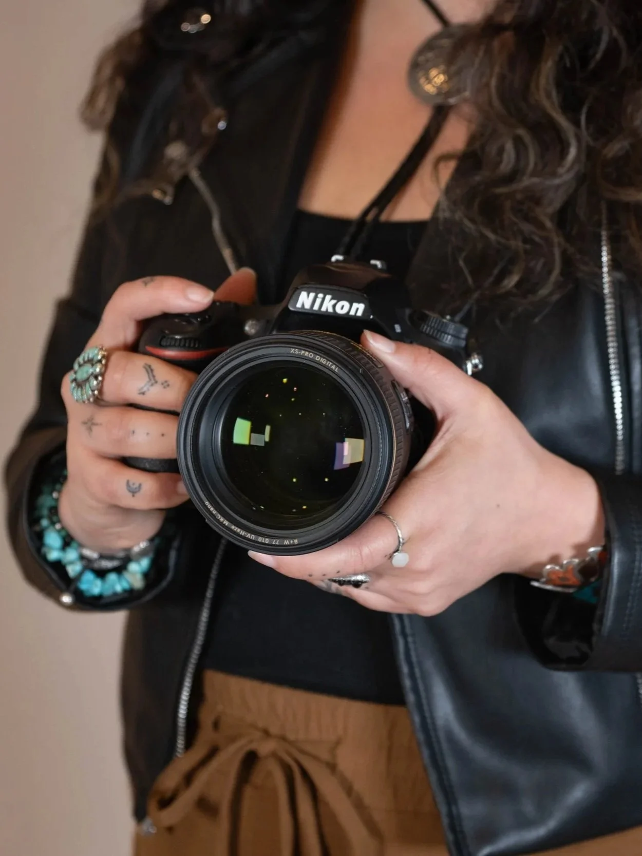 A woman taking a selfie with a Nikon camera, wearing a black leather jacket, turquoise jewelry, and multiple rings. She has wavy hair and is standing indoors.
