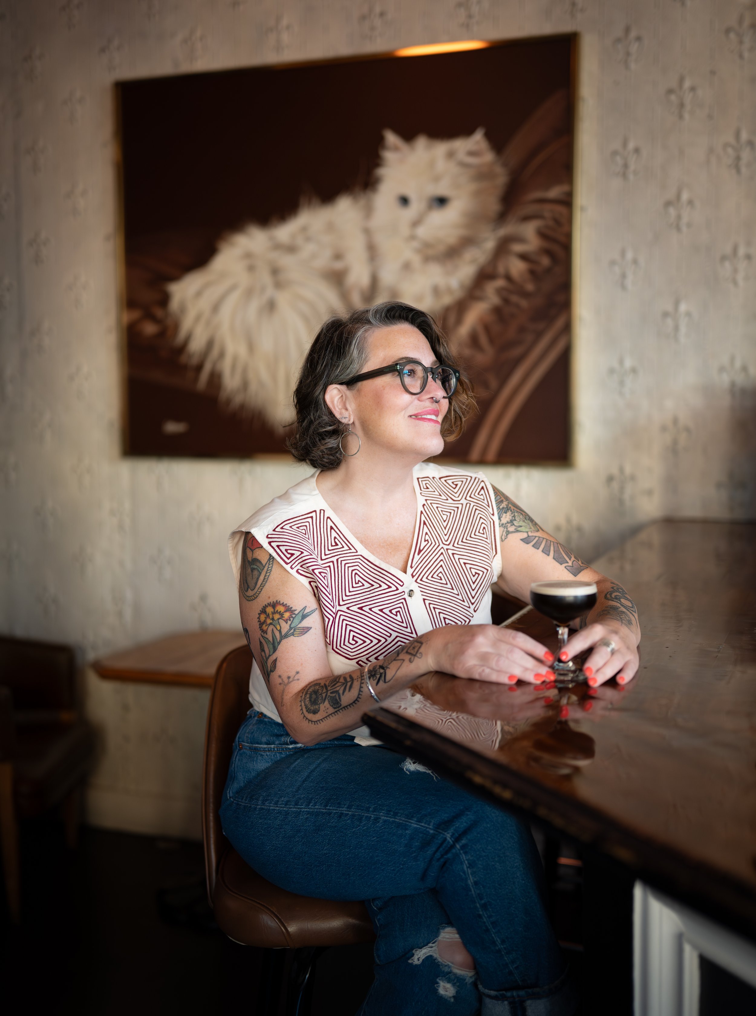A woman with dark wavy hair, wearing glasses, a sleeveless patterned top, and ripped jeans, sitting at a bar with a cocktail glass in her hands. Behind her hangs a portrait of a fluffy white cat with blue eyes.