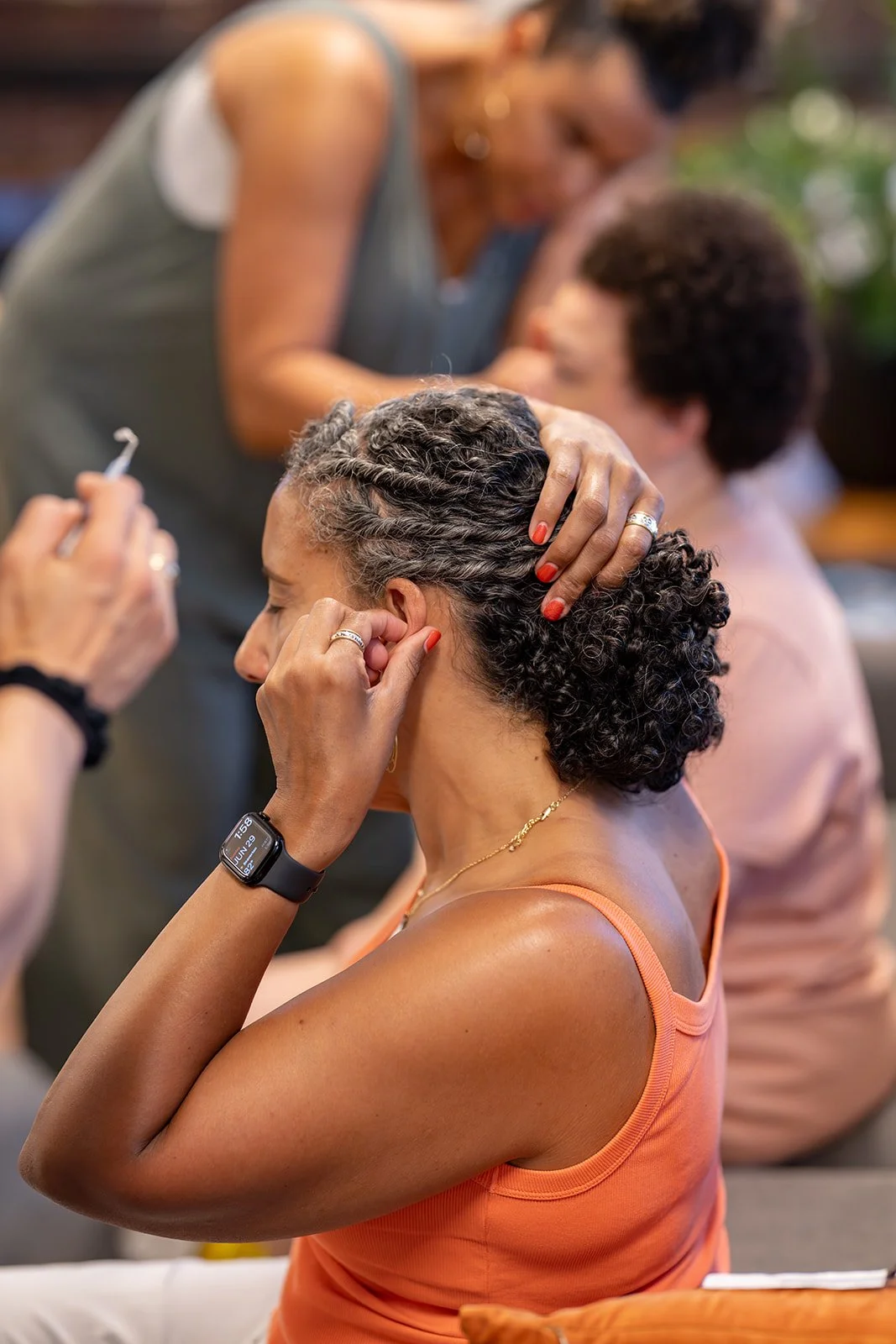A woman with short, curly hair getting her ear pierced by a professional, with another woman in the background.