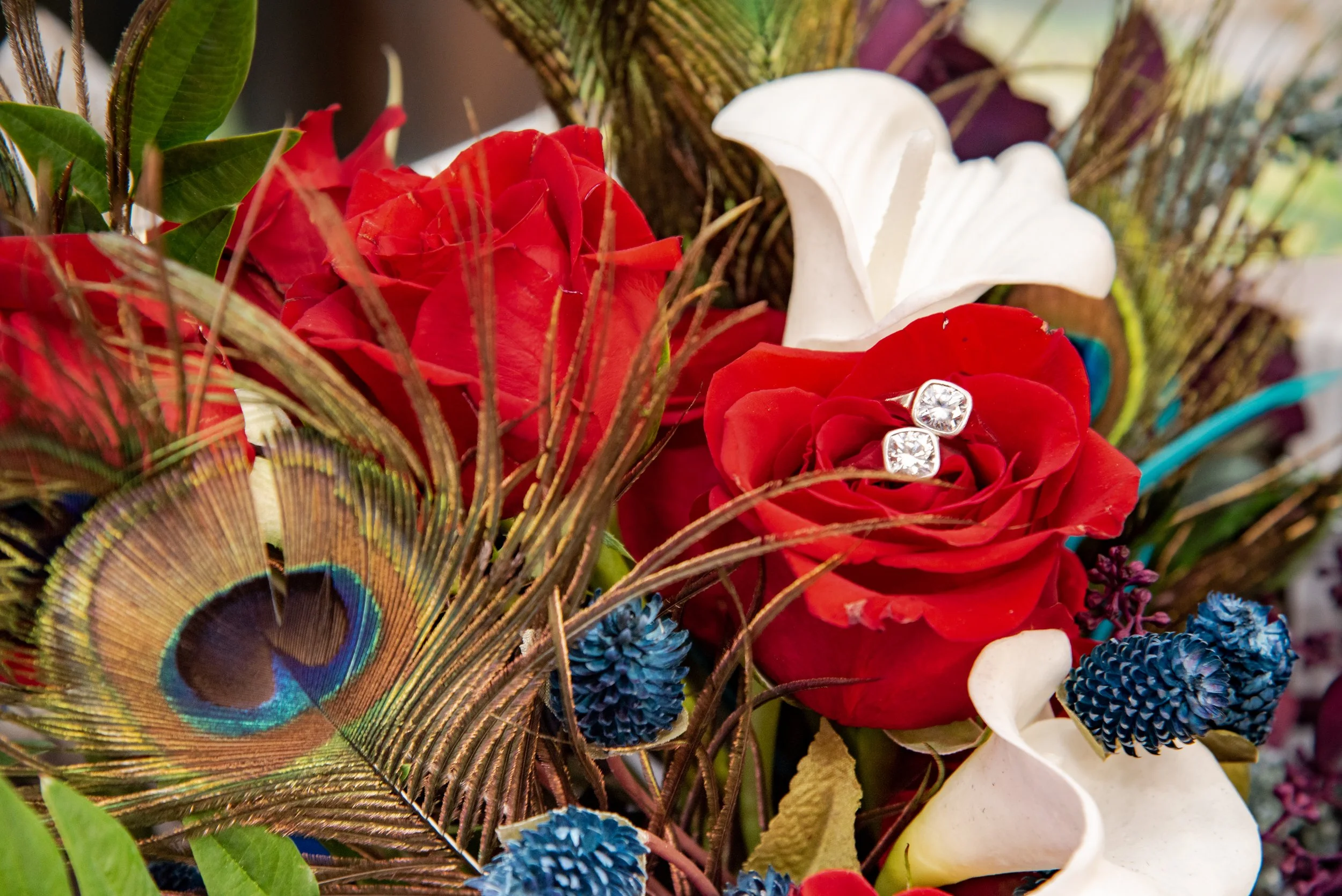 A bouquet of flowers with red roses, white lilies, blue flowers, peacock feathers, and gemstone earrings.