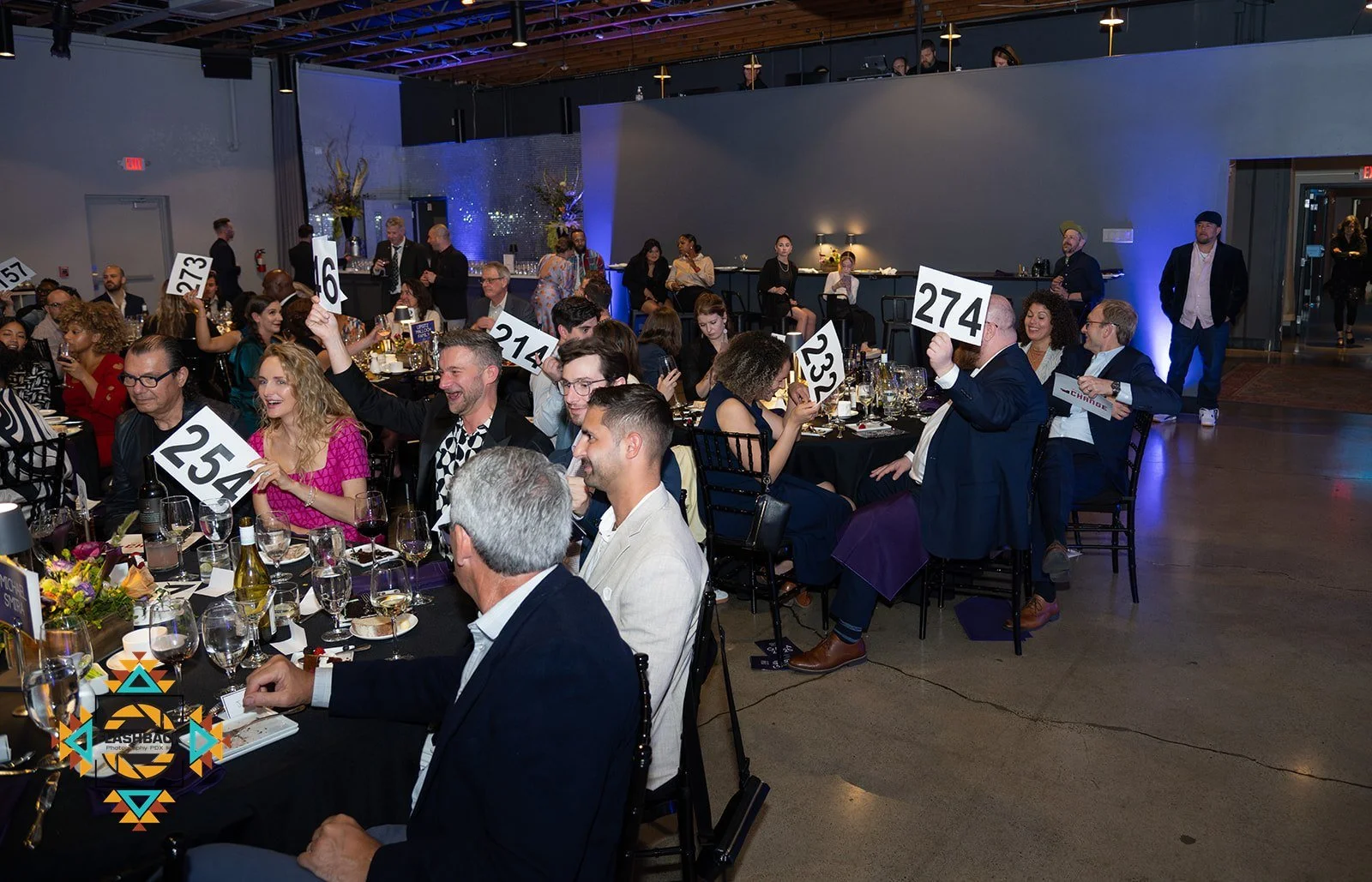 People attending a silent auction at a formal event, seated at tables with wine glasses, and holding up bid numbers.