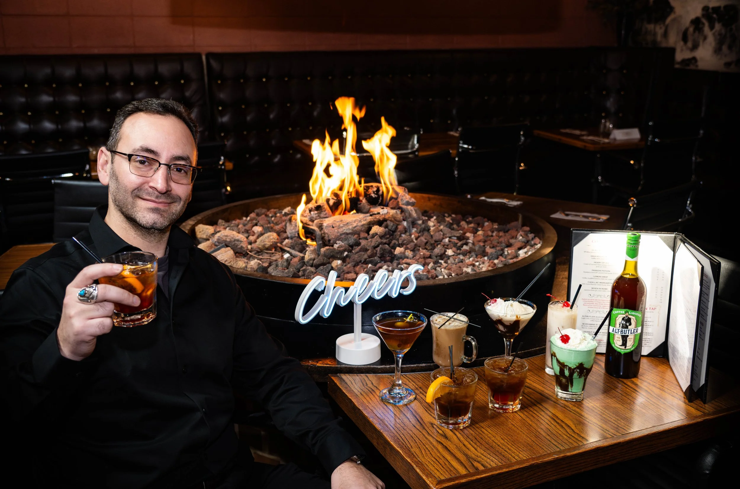 A man in black shirt with glasses holding a drink while sitting at a table with various cocktails, a neon 'Cheers' sign, and a flame in a fire pit in the background inside a restaurant.