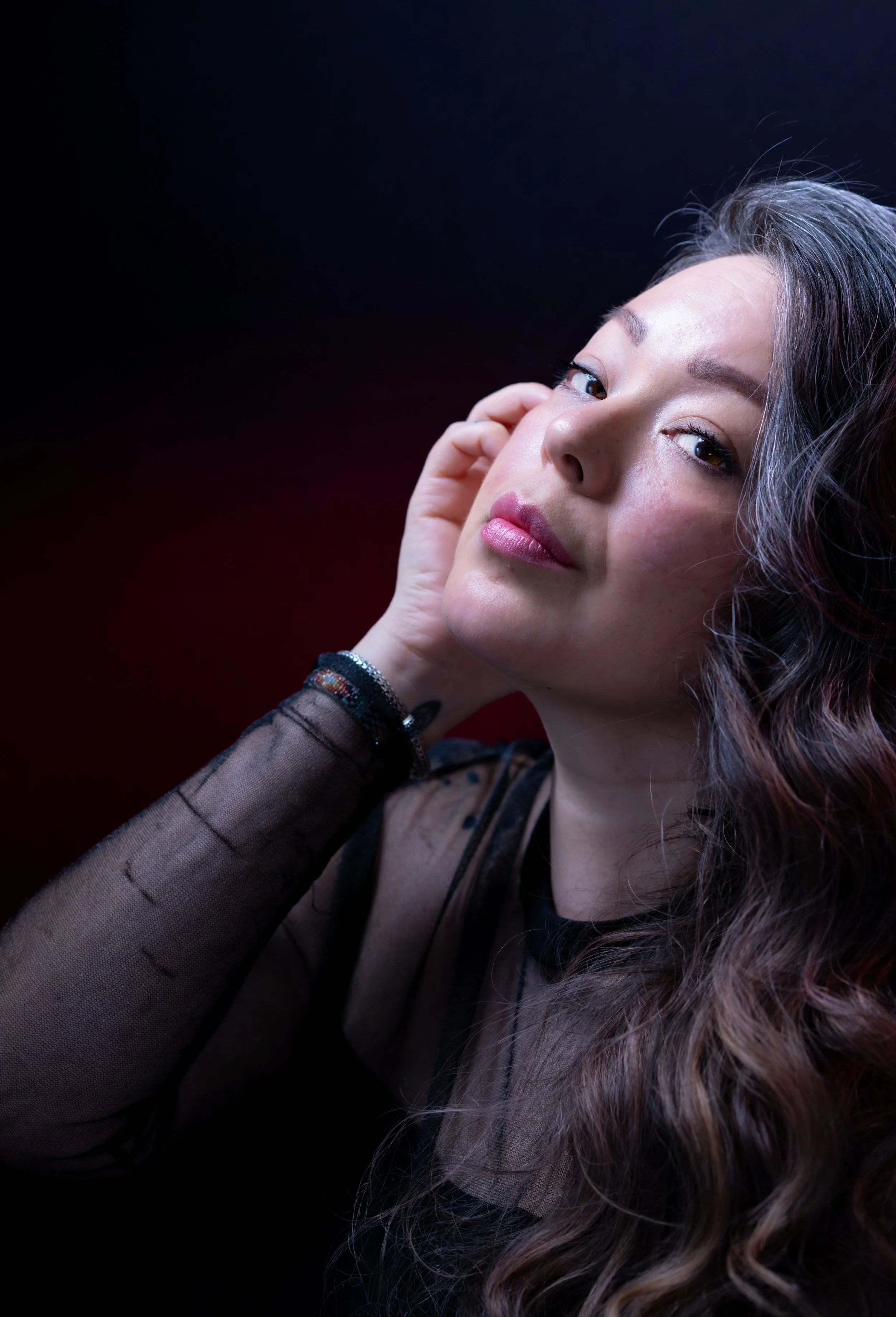 Portrait of a woman with long wavy hair, wearing a black, sheer top, touching her face with her left hand, against a dark red background.