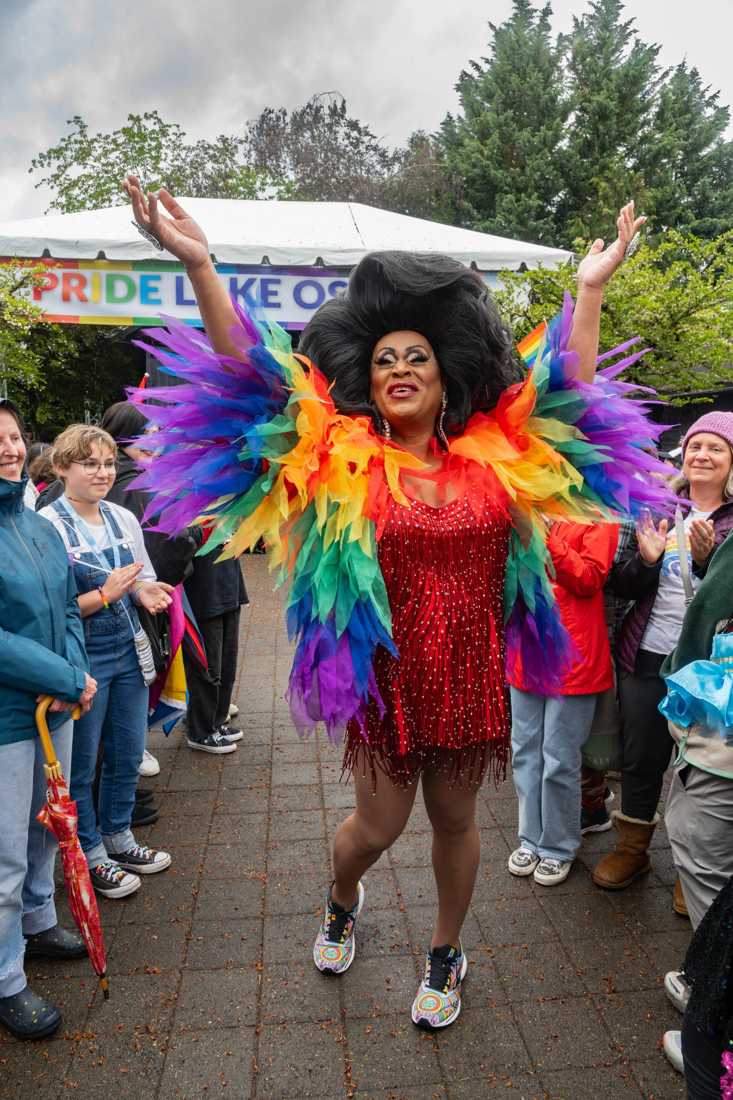 A drag performer dressed in a rainbow feathered costume and red sequined dress, dancing at a pride event with a diverse crowd celebrating and a banner that says 'PRIDE LIKE OS' in the background.