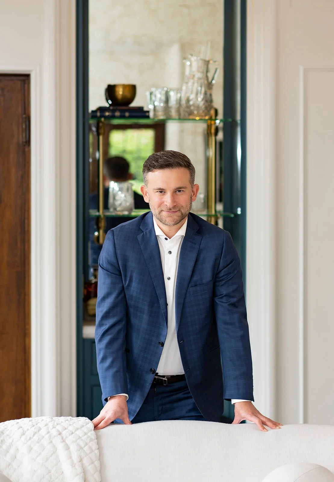 A man in a navy suit and white shirt leaning on the back of a white sofa in a well-decorated living room.
