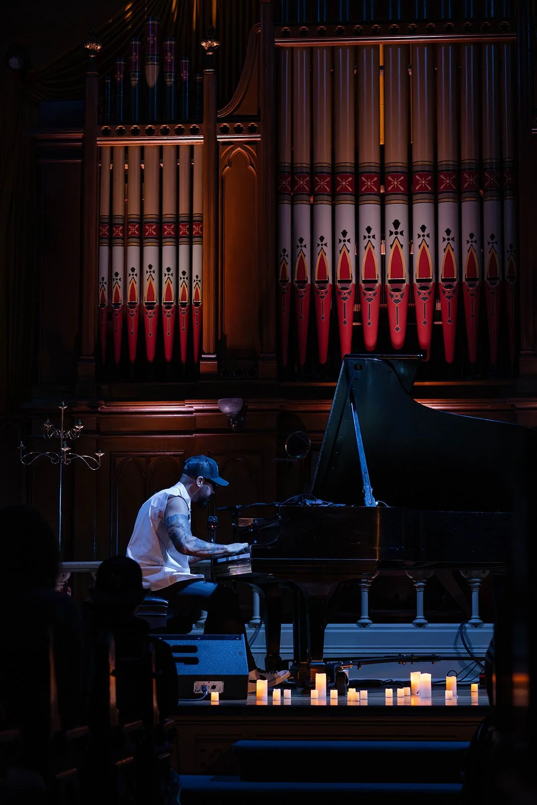 A musician playing a grand piano on stage decorated with lit candles, large organ pipes in the background, and an audience in the foreground.