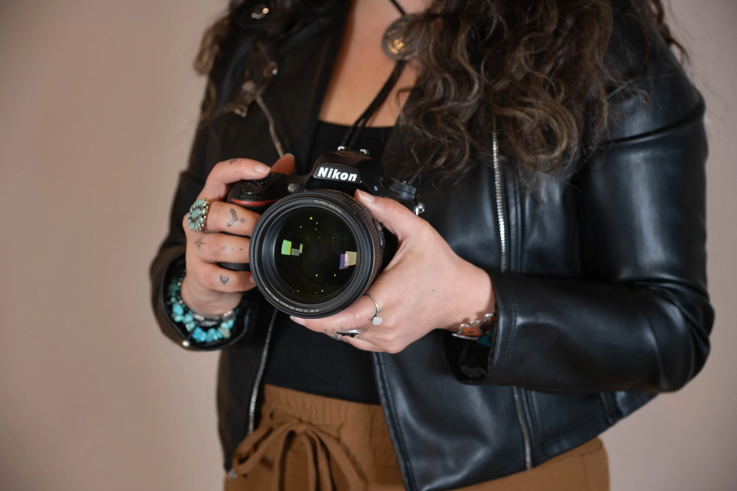 Person holding a Nikon camera, wearing a black leather jacket, with visible rings and turquoise bracelets.