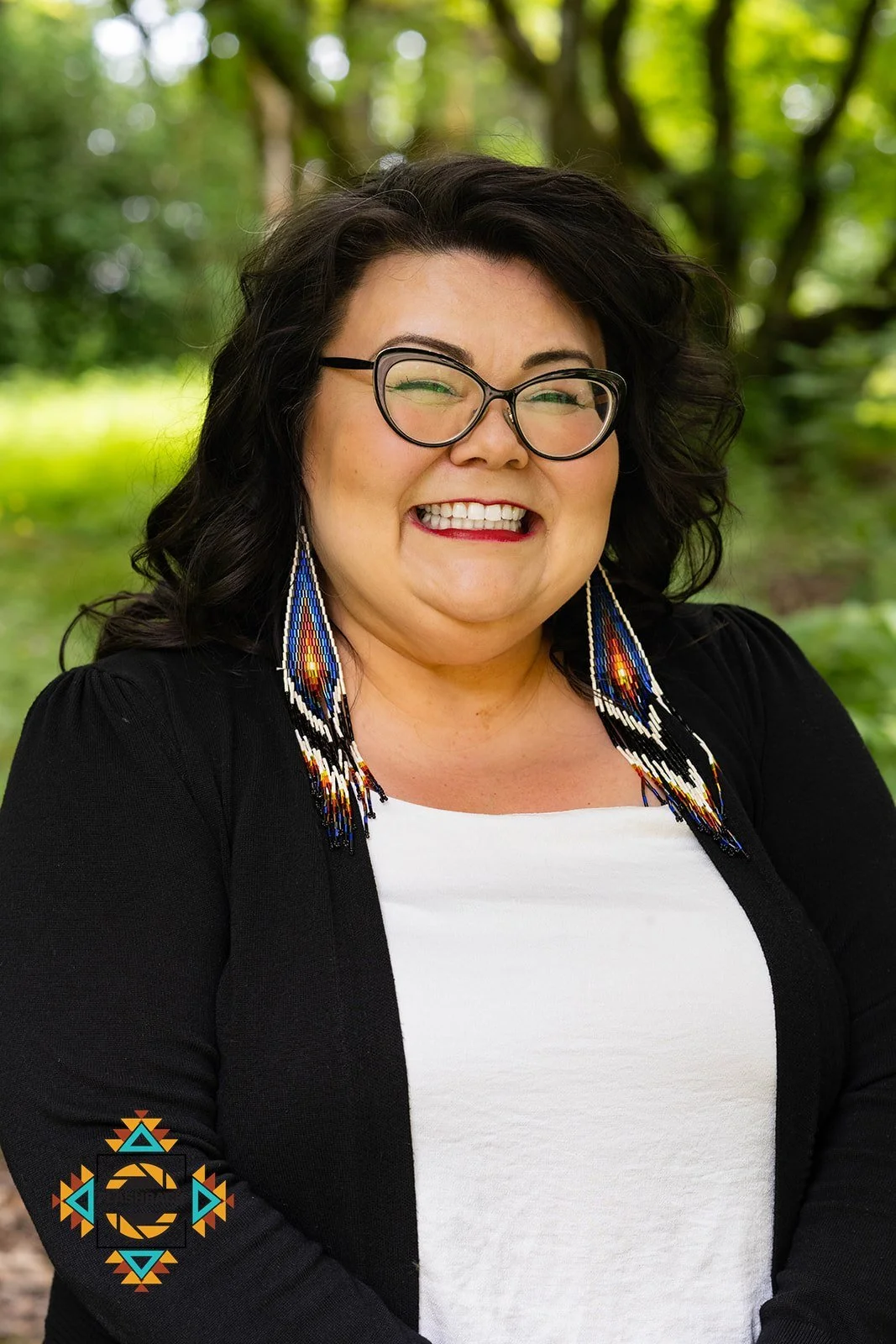 Smiling woman with dark curly hair and glasses, wearing beaded earrings, a white top, and a black jacket, standing outdoors with green trees in the background.