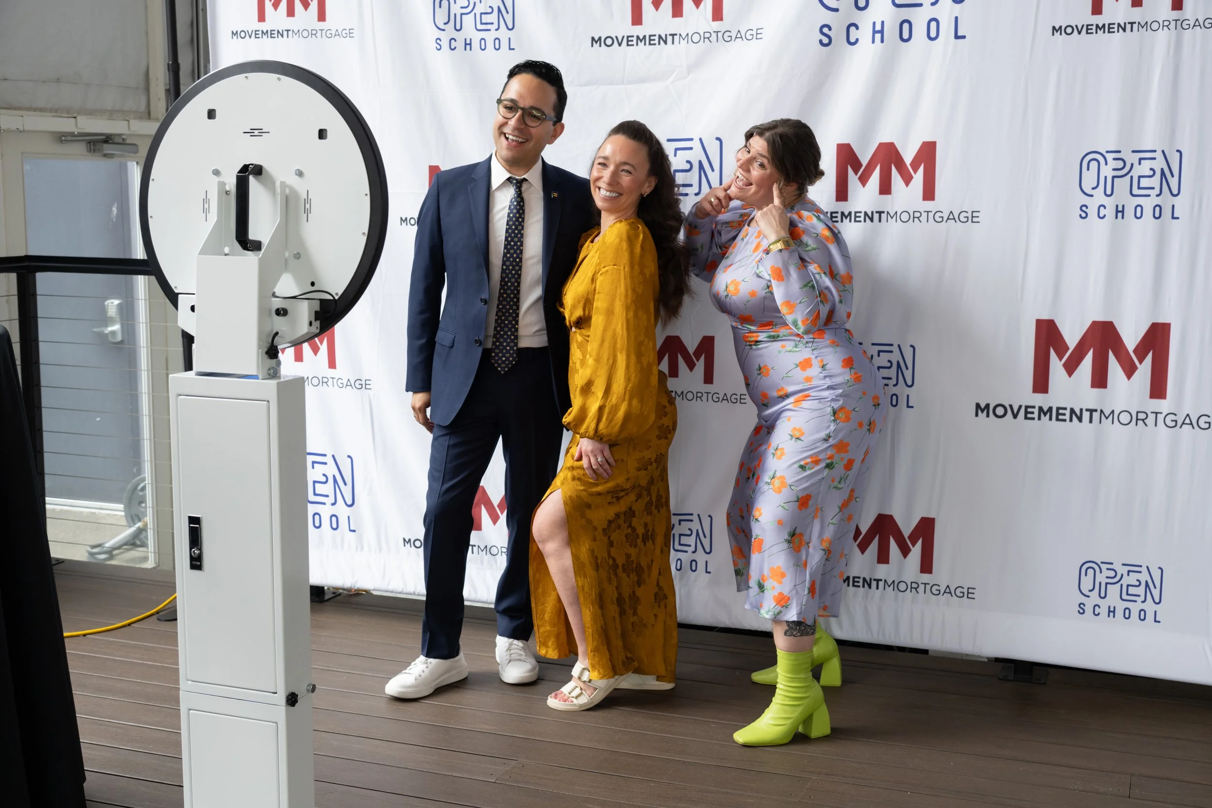 Three people posing for a photo in front of a backdrop with logos. The person on the left is a man in a navy suit with glasses, the woman in the middle is wearing a yellow dress, and the woman on the right is in a floral jumpsuit with bright green boots. They are smiling and standing next to a large machine.