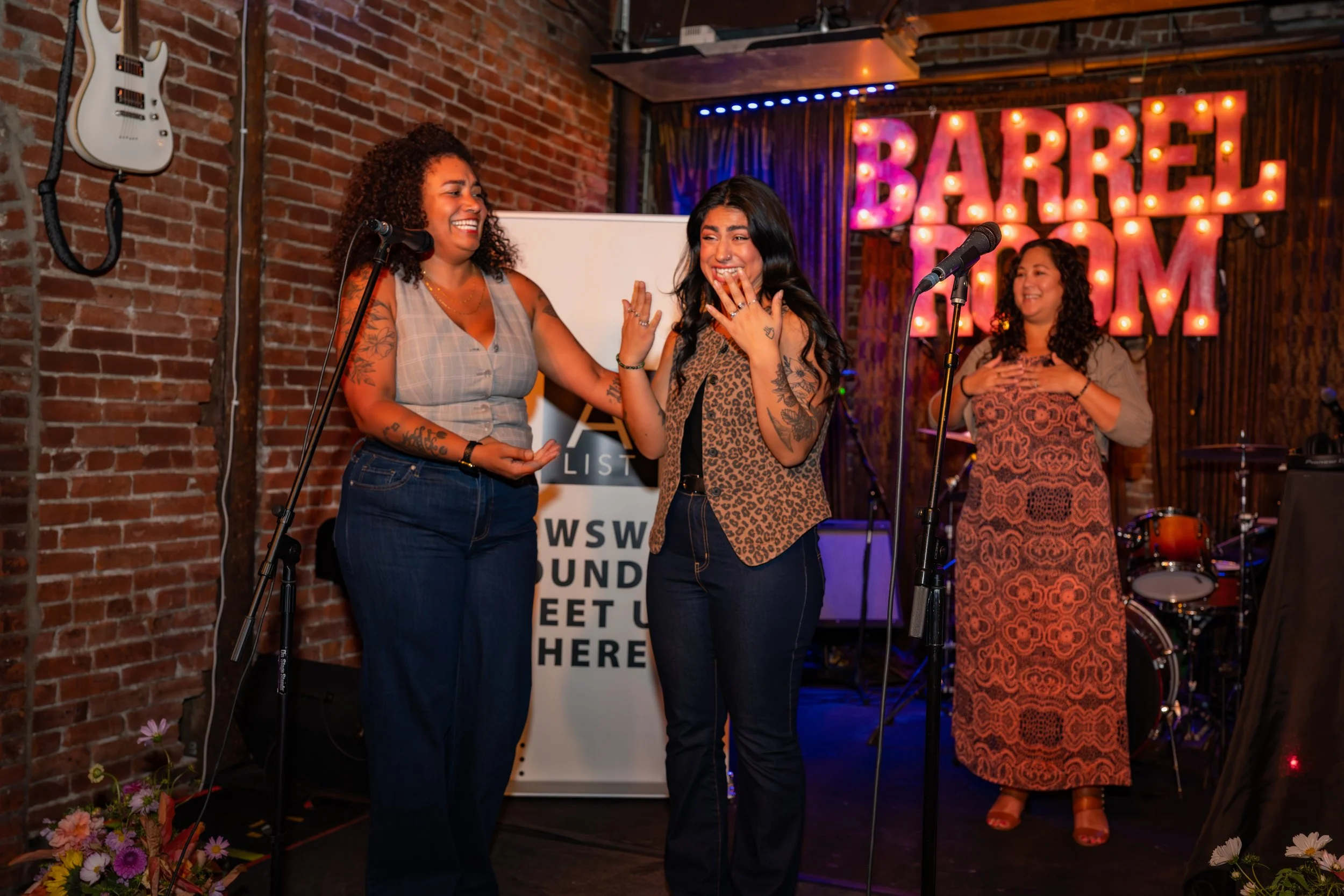 Three women on stage at Barrel Room, two in the front laughing and talking, one in the back with hands on chest, surrounded by musical instruments and stage lights.