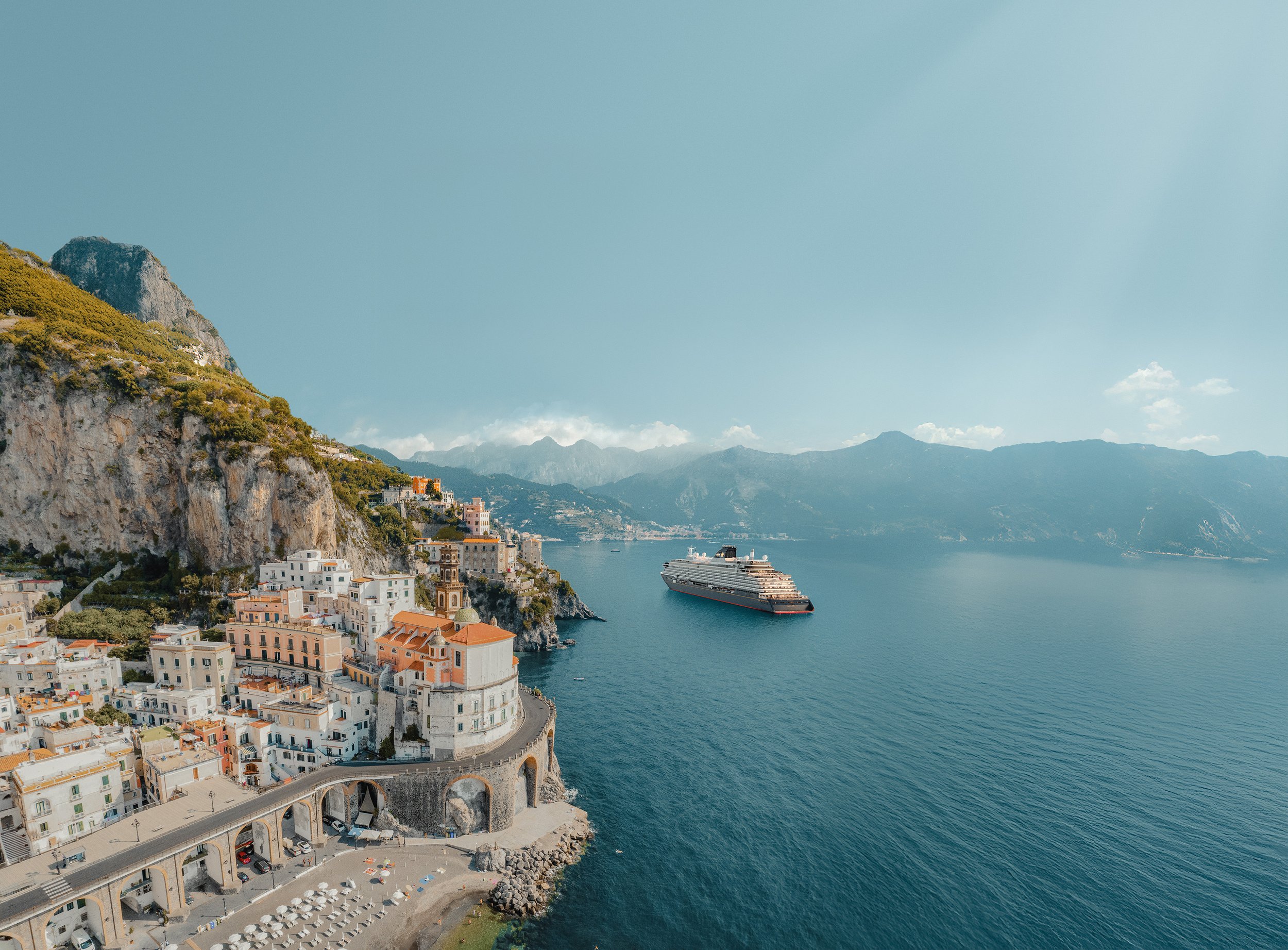 Coastal town with colorful buildings on a hillside next to a large body of water, a cruise ship anchored in the water, mountains in the distance, and a clear sky.