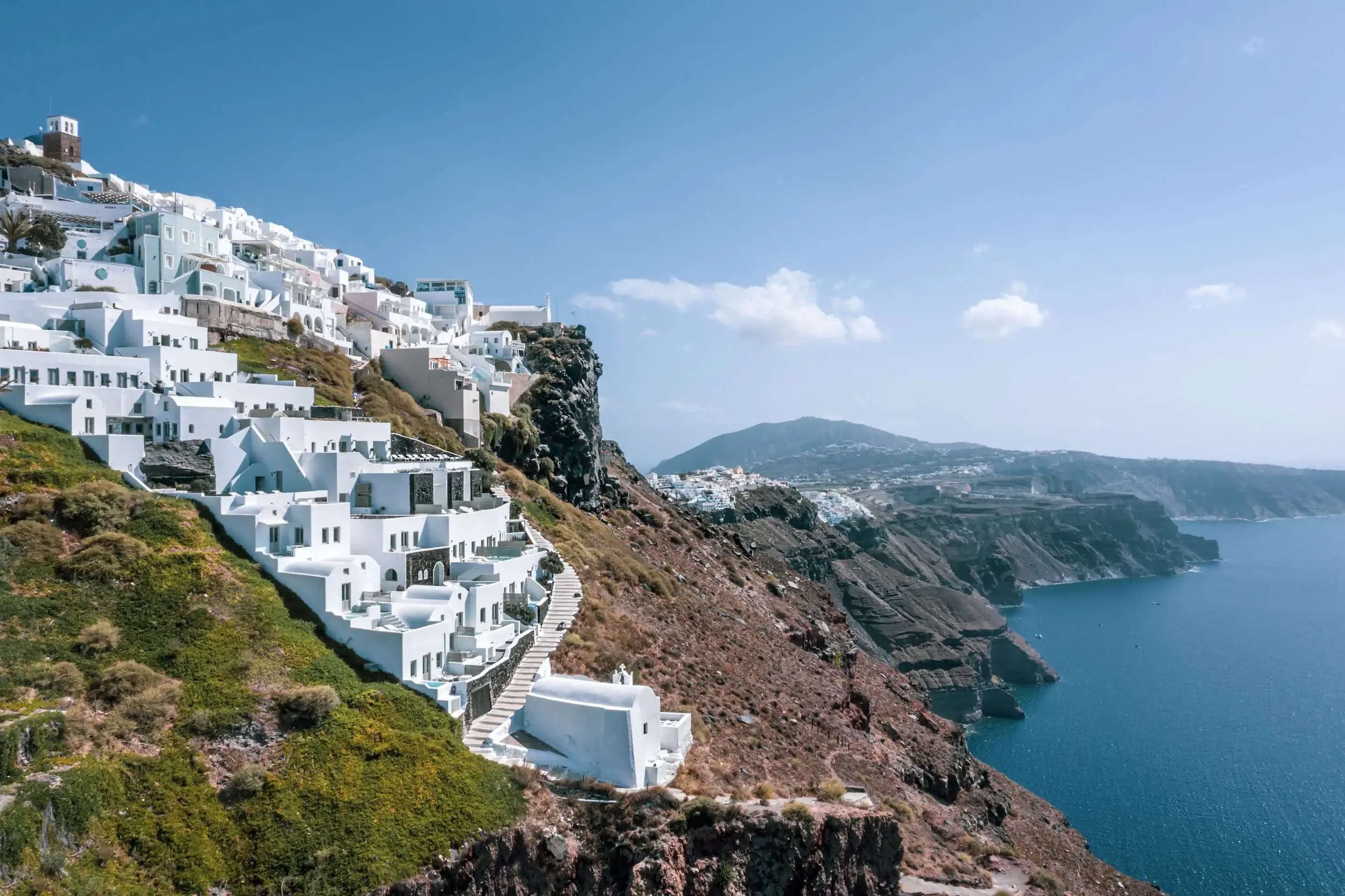 View of white buildings with blue accents on a cliffside overlooking the blue ocean in Santorini, Greece.