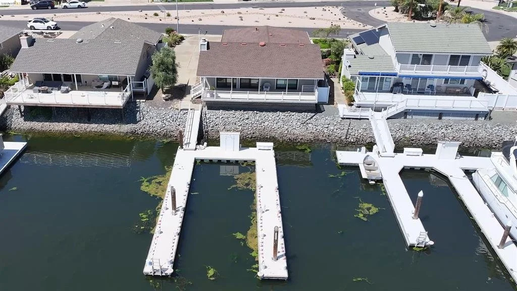 From an aerial view, three houses with a water frontage and docks are visible. The rightmost house has a staircase leading down to the dock, with boats moored alongside. The middle house has a similar dock, but no boat is visible. The left house has a large deck overlooking the water. The houses are separated by rocks and a seawall, with a road running behind them.