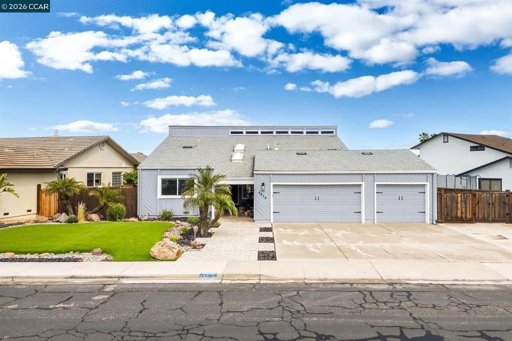 A modern house with a gray exterior, three garage doors, and a front yard with a small palm tree, artificial grass, and rocks, under a partly cloudy blue sky.