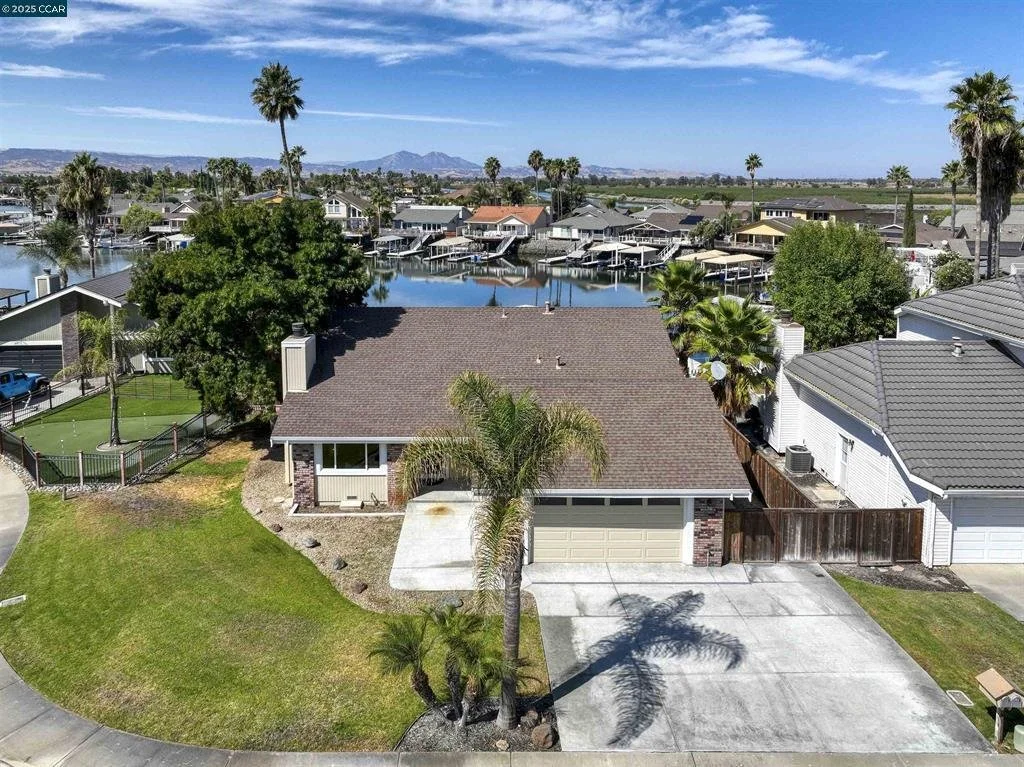 A house with a brown roof and attached garage in a neighborhood with palm trees, next to a waterway with docks, under a blue sky with scattered clouds.