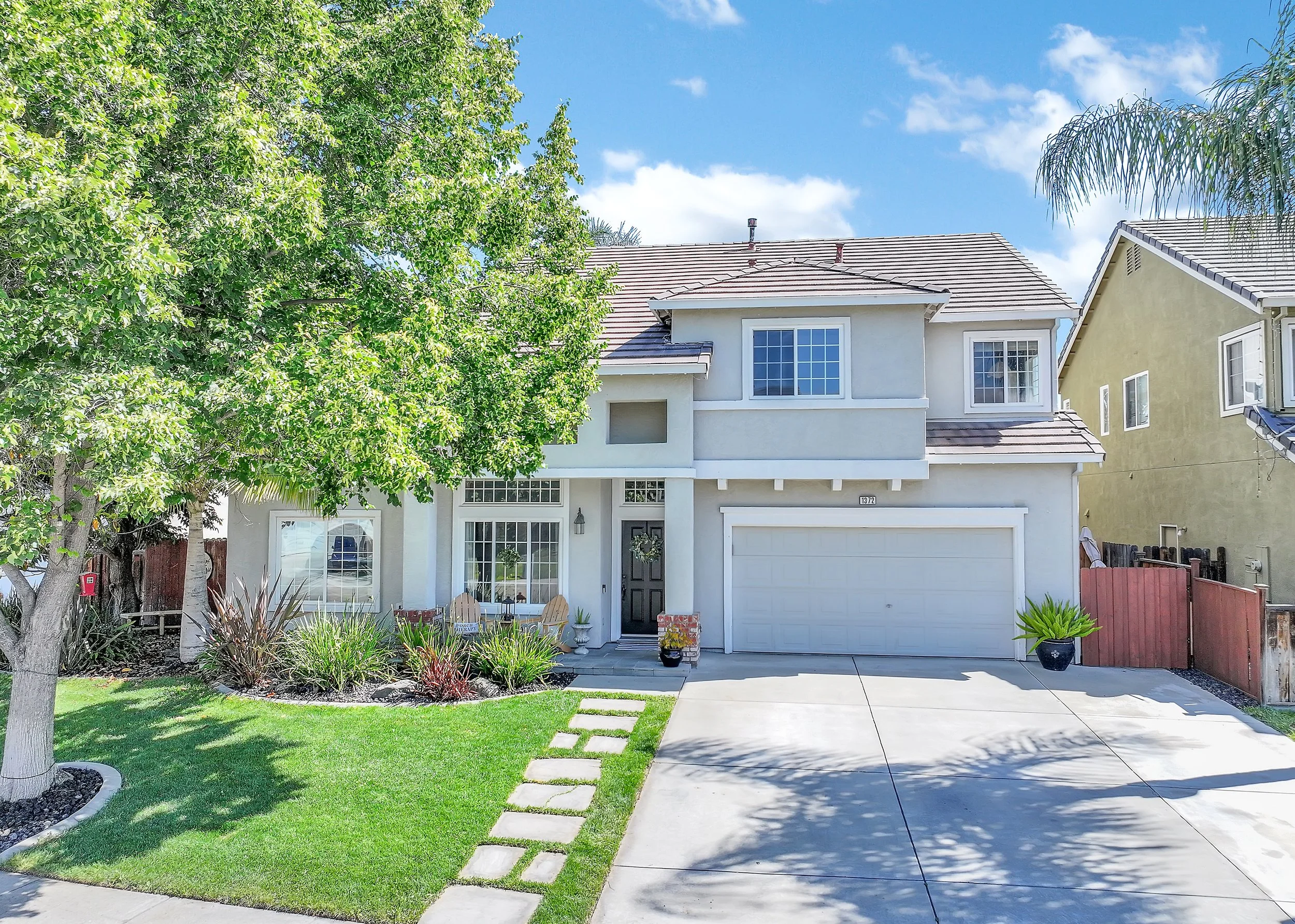 A white two-story house with a gray tiled roof, front door, and attached garage, surrounded by a well-maintained lawn, a sidewalk with stepping stones, and various plants and trees in a suburban neighborhood under a bright blue sky.