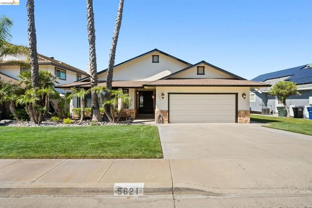 A single-story suburban house with a front lawn, palm trees, and a driveway leading to a two-car garage. The house has beige siding with stone accents at the bottom and a dark-colored front door.