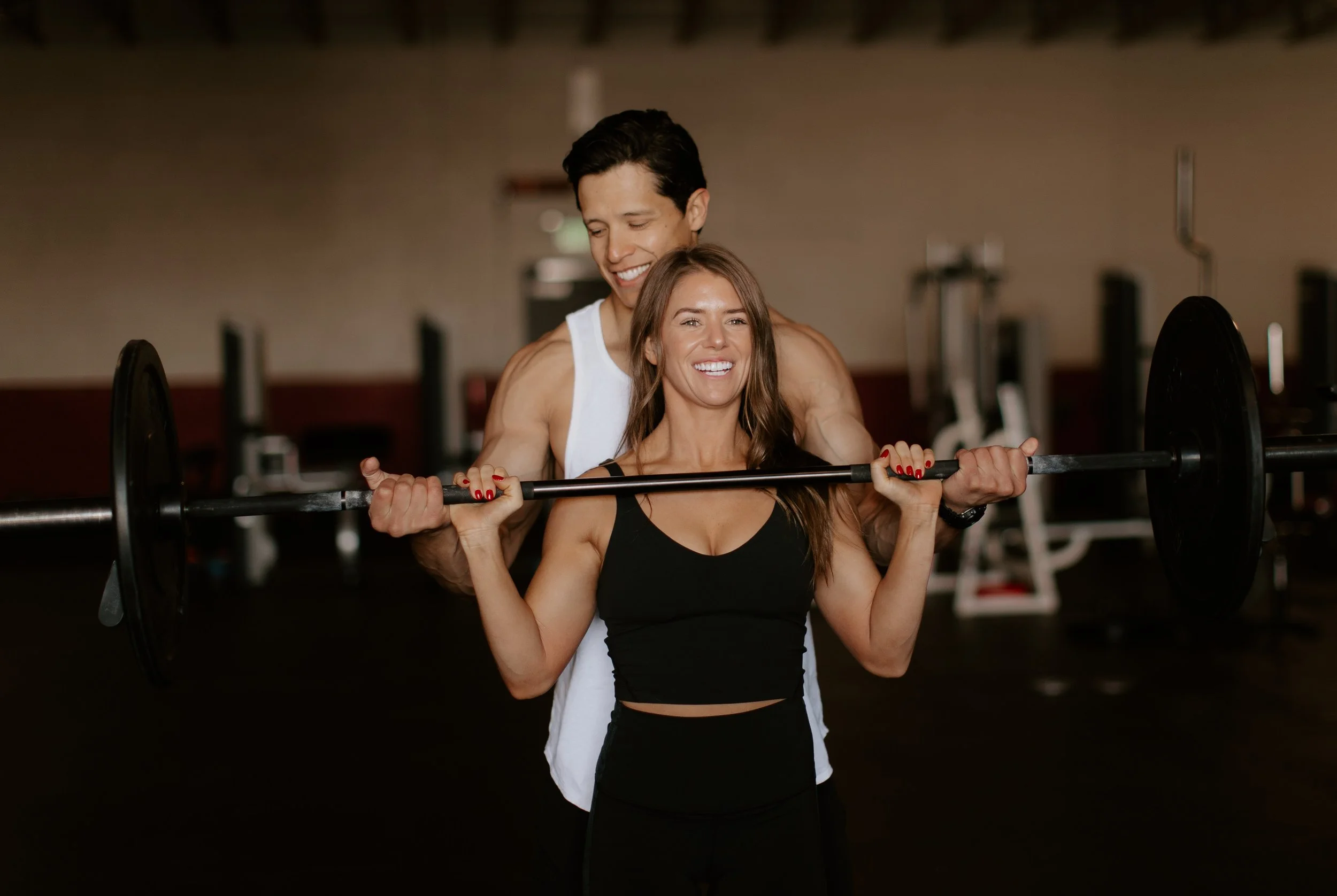 A man and woman training together at the gym, with the woman lifting a barbell and the man assisting or spotting her, both smiling.