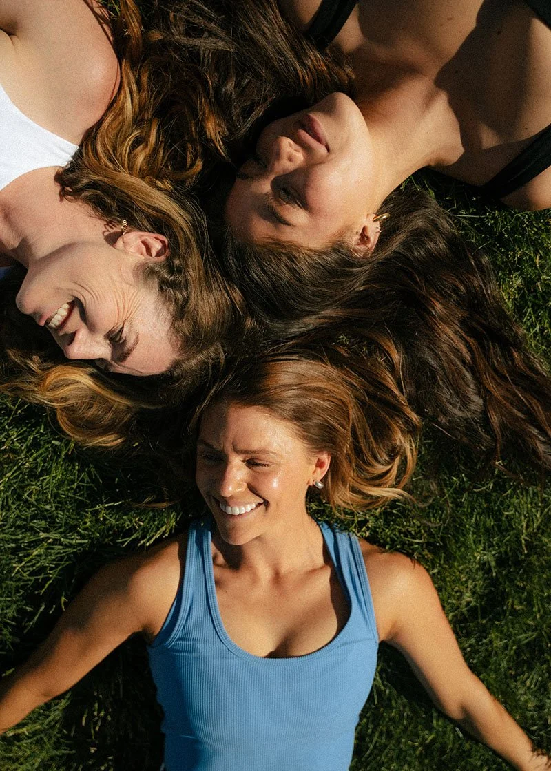 Four women lying on grass with heads together, smiling and laughing.