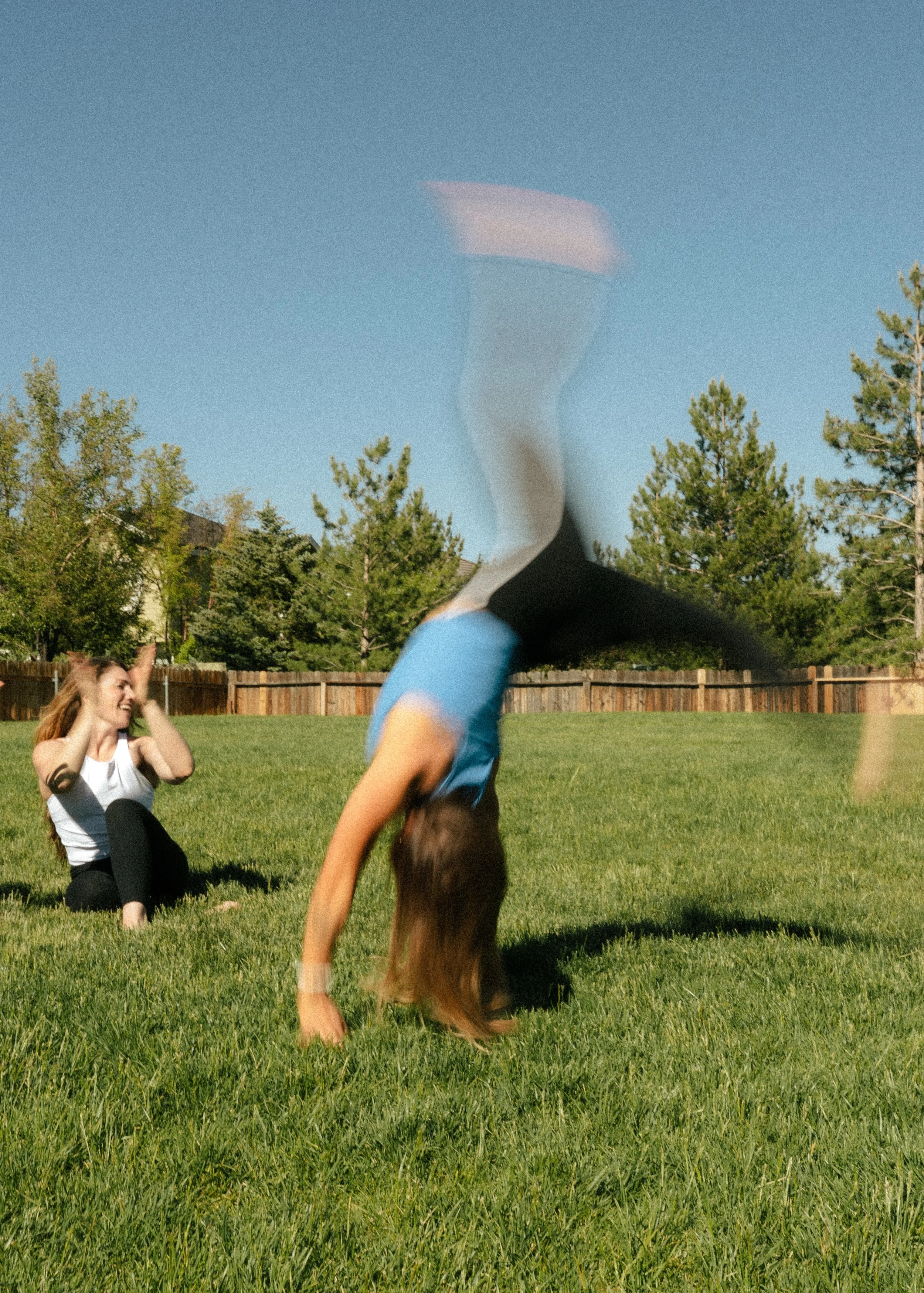 Two women playing in a backyard on a sunny day, one doing a handstand and the other sitting on the grass laughing.