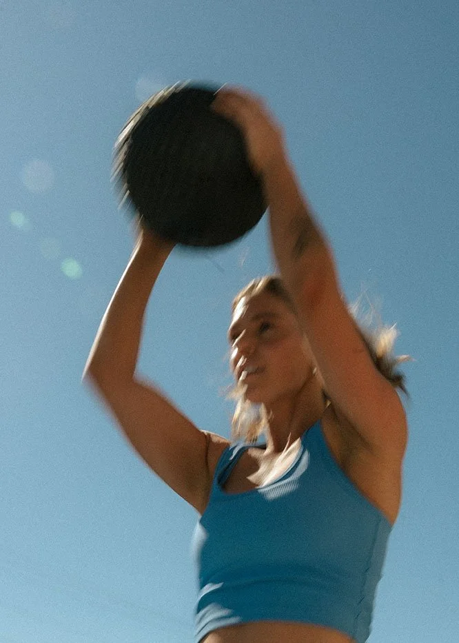 A woman in athletic wear preparing to throw a medicine ball outdoors under a clear blue sky.