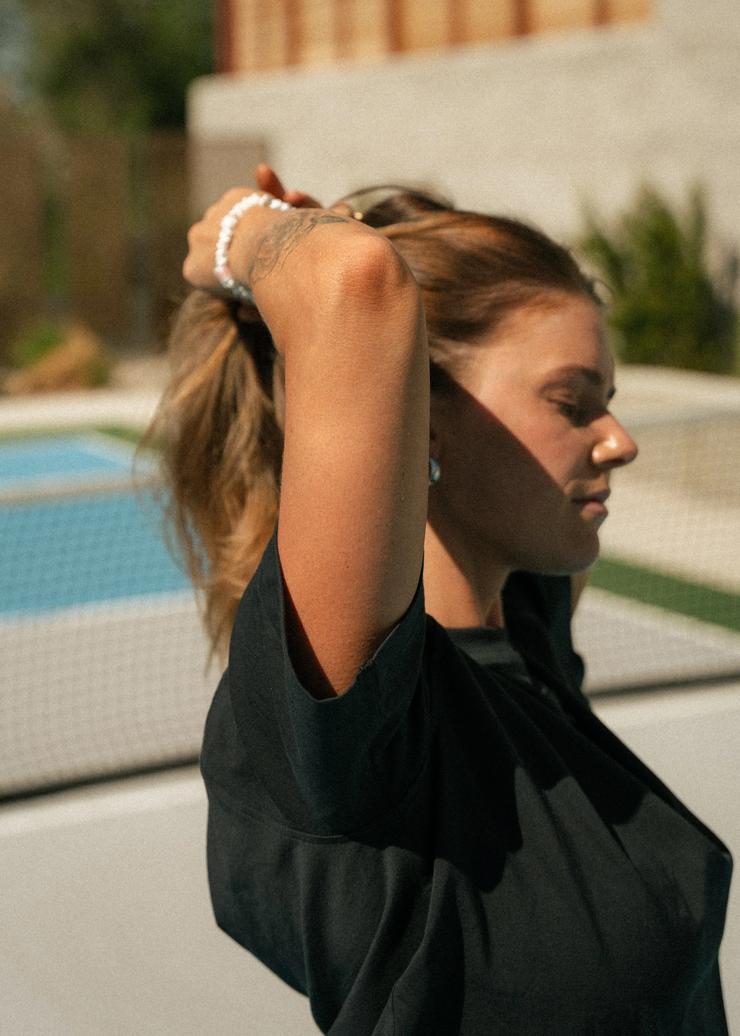 A woman with light brown hair and a tattoo on her arm, wearing a black shirt, standing outdoors with her eyes closed and hands behind her head.