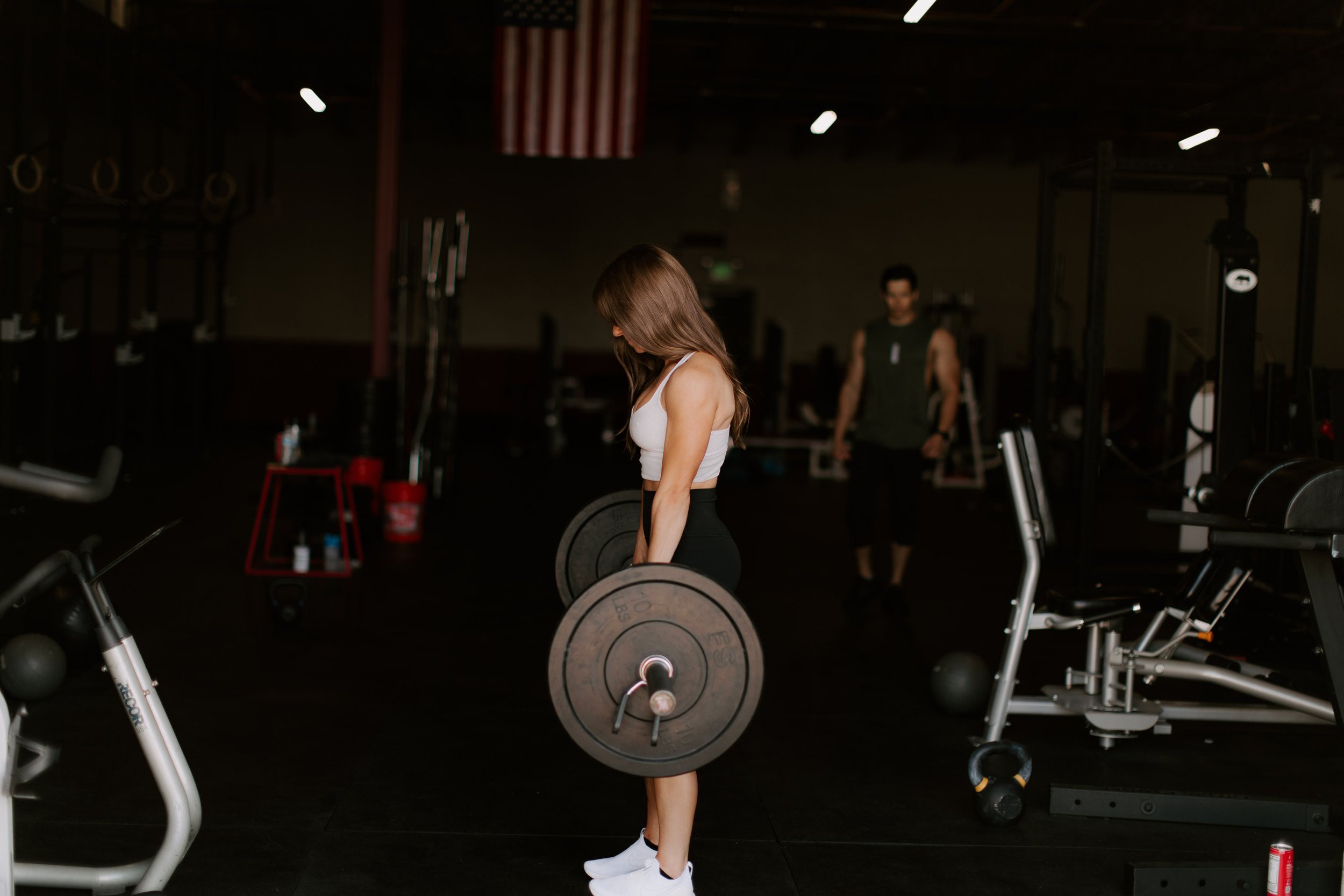 Woman in workout clothes lifting a barbell with weights at the gym, with a man in the background and gym equipment around.