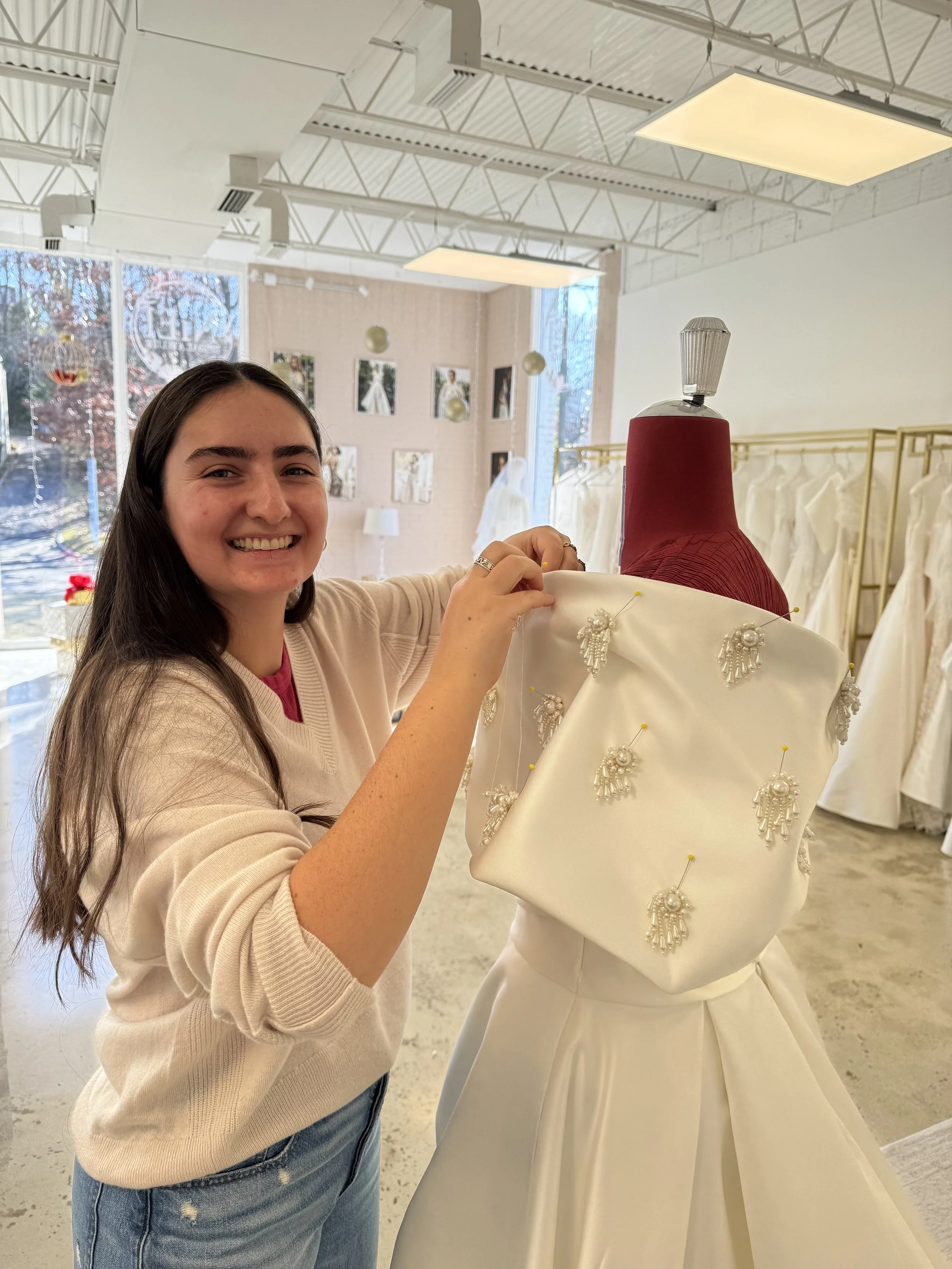 Young woman smiling in a bridal boutique, standing next to a mannequin wearing a wedding dress adorned with pearl and beaded embellishments.