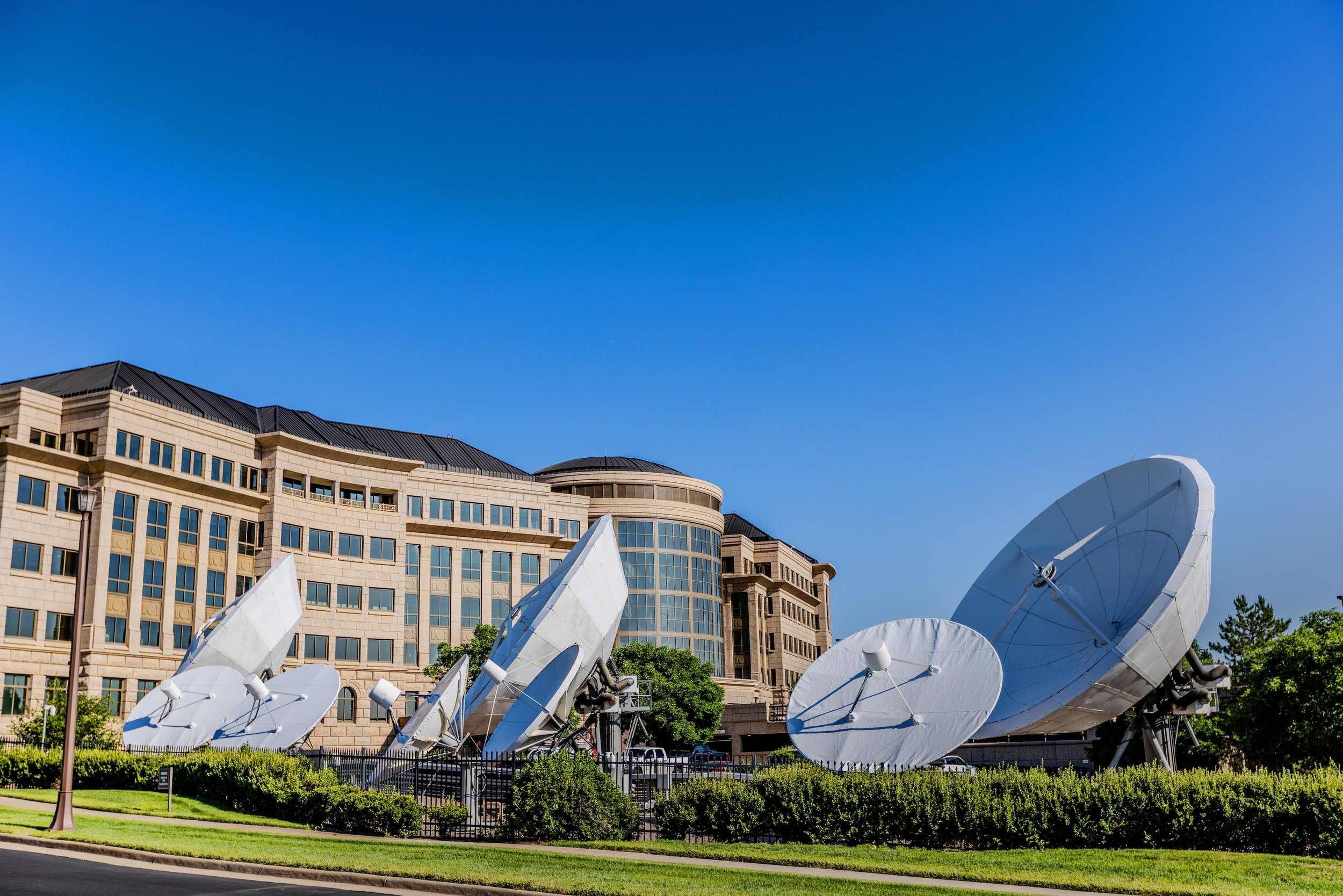 Multiple satellite dishes in front of a large beige and brown building under a clear blue sky.