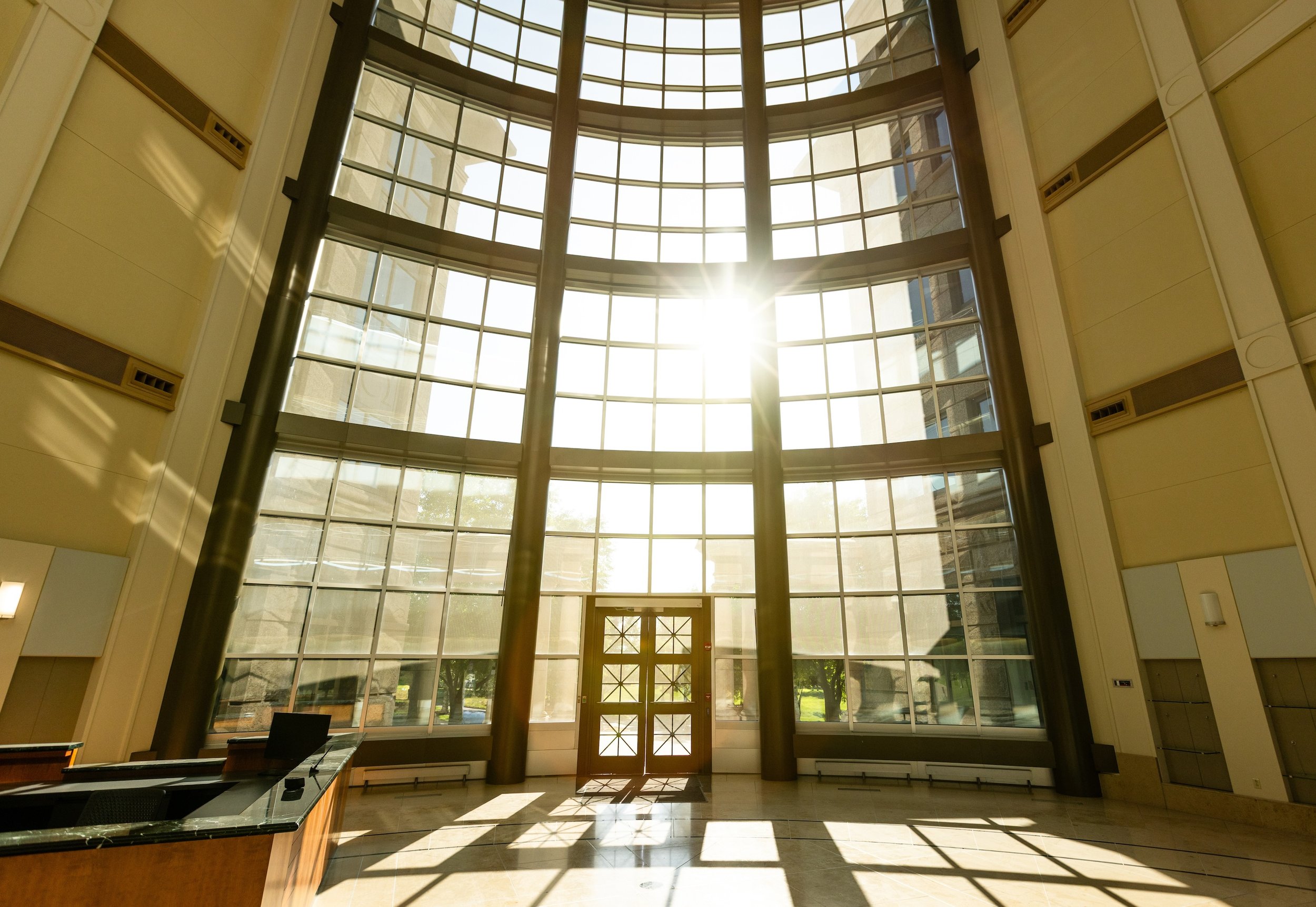 Interior of a modern building with large glass windows and sunlight streaming through, casting shadows on the floor near a central door.