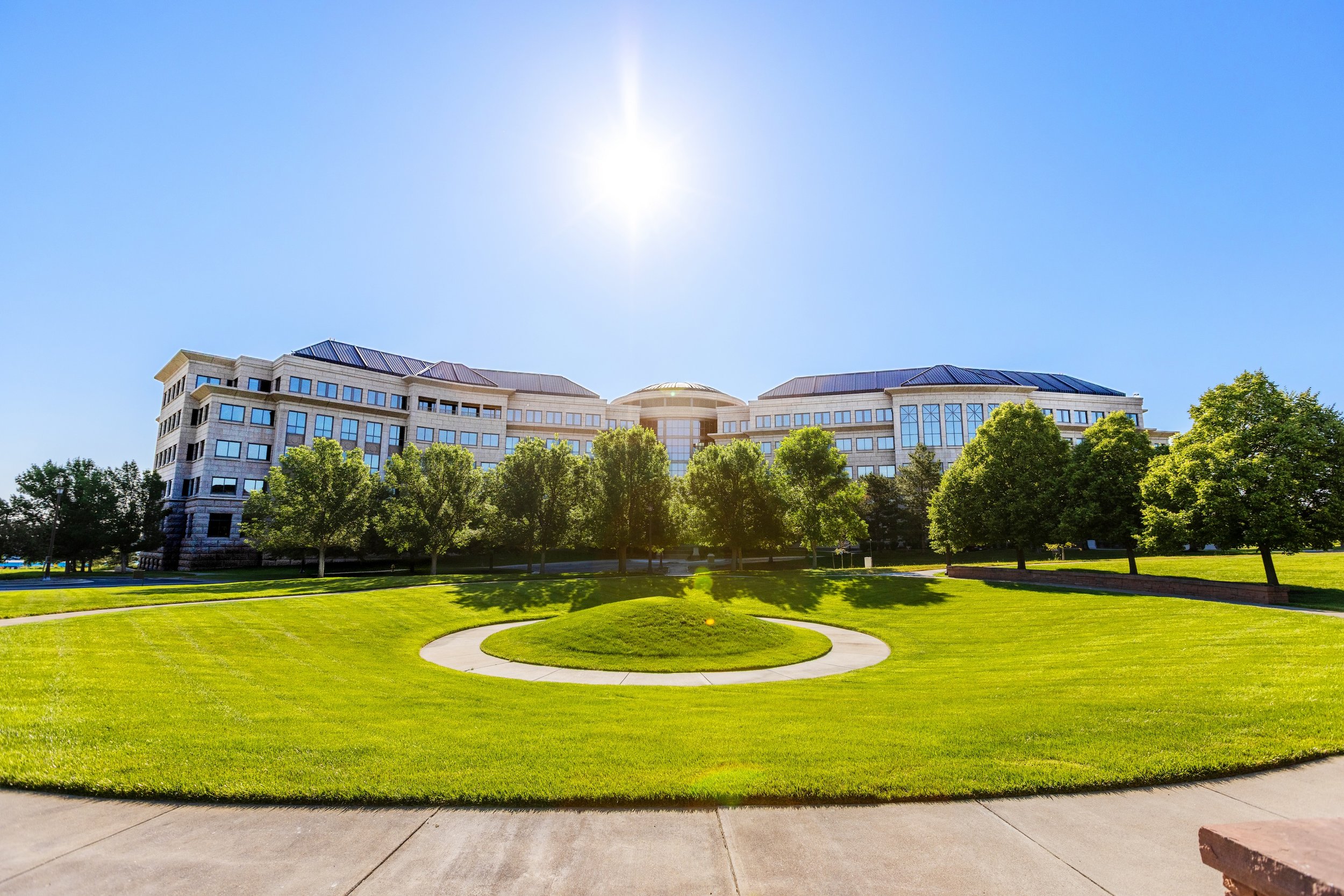 A large multi-story building with modern architecture, surrounded by green trees and a well-maintained lawn, with the sun shining brightly in a clear blue sky.