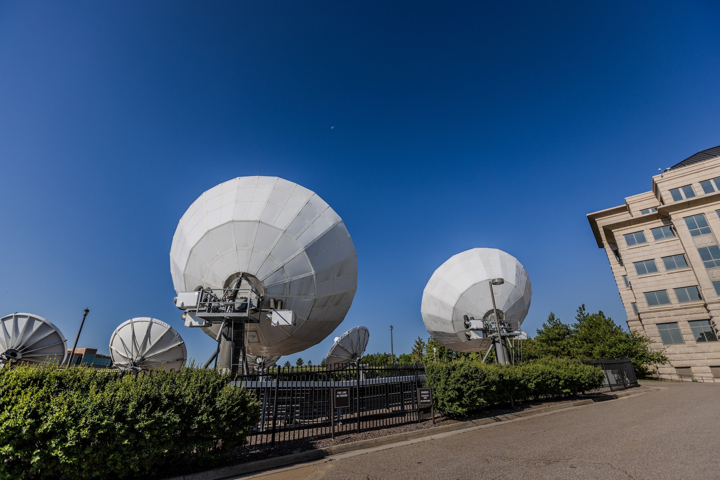 Multiple large satellite dishes outdoors, with a clear blue sky and a building in the background.