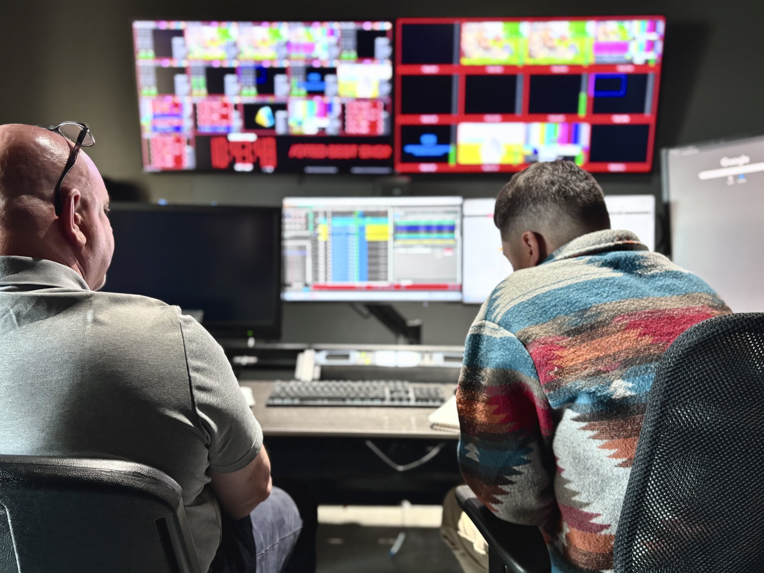 Two men are working at a control room with multiple monitors displaying colorful technical data, schedules, and program feeds.