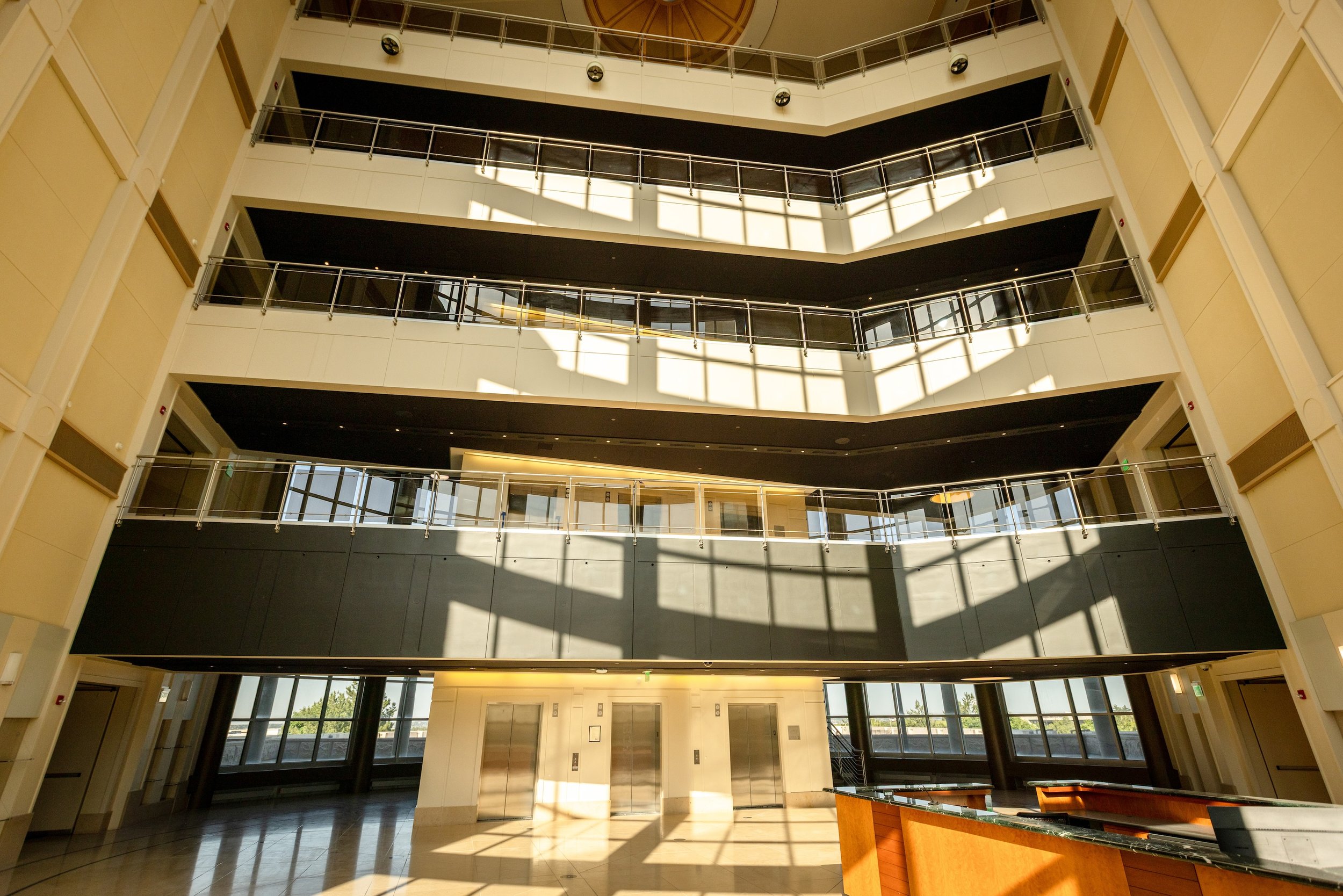 Interior of a modern multi-story building with a lobby, elevators, and balconies, with sunlight streaming through large windows.