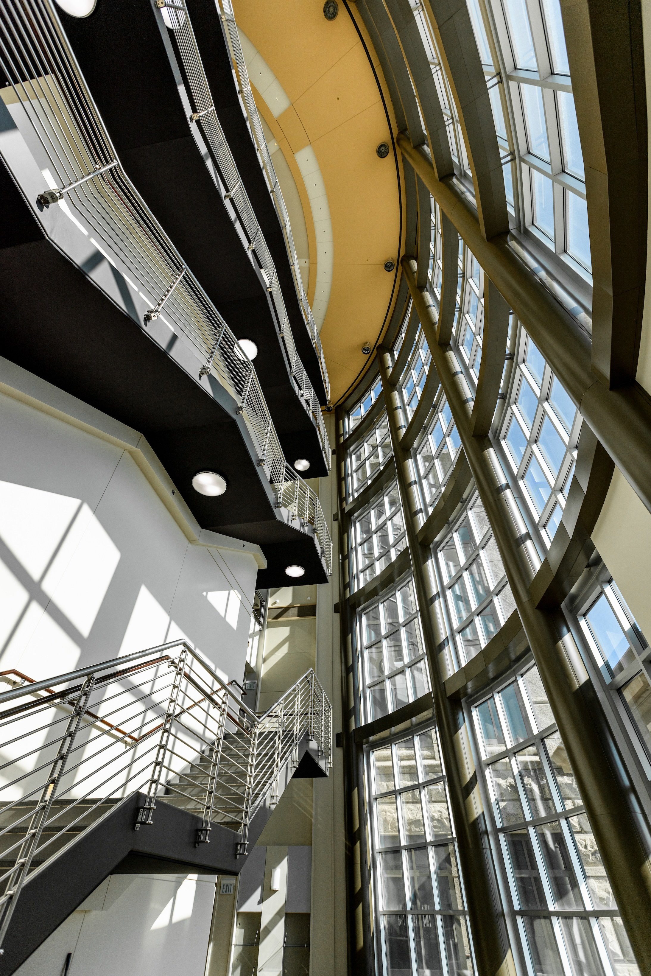 Interior view of a modern building with multiple levels, glass windows, and metal railings, with sunlight casting shadows on the walls.