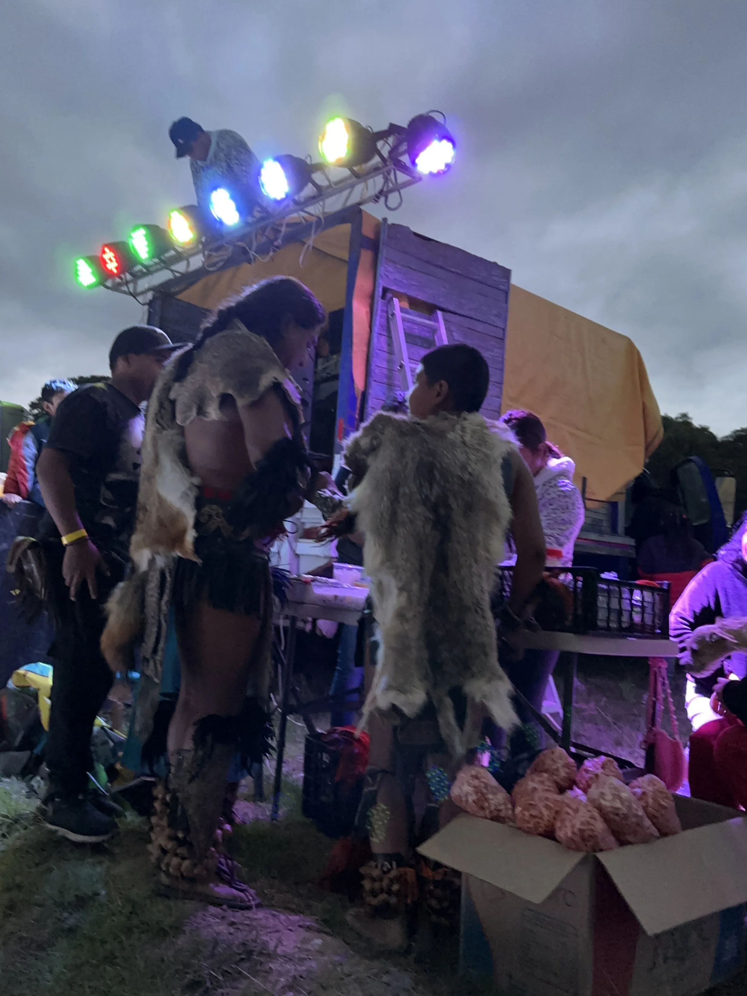 Native American clothing at an outdoor event during dusk, with a decorated wooden stage and colorful lights.part our rituals at camp tlalankann la pedrera tlalancaleca