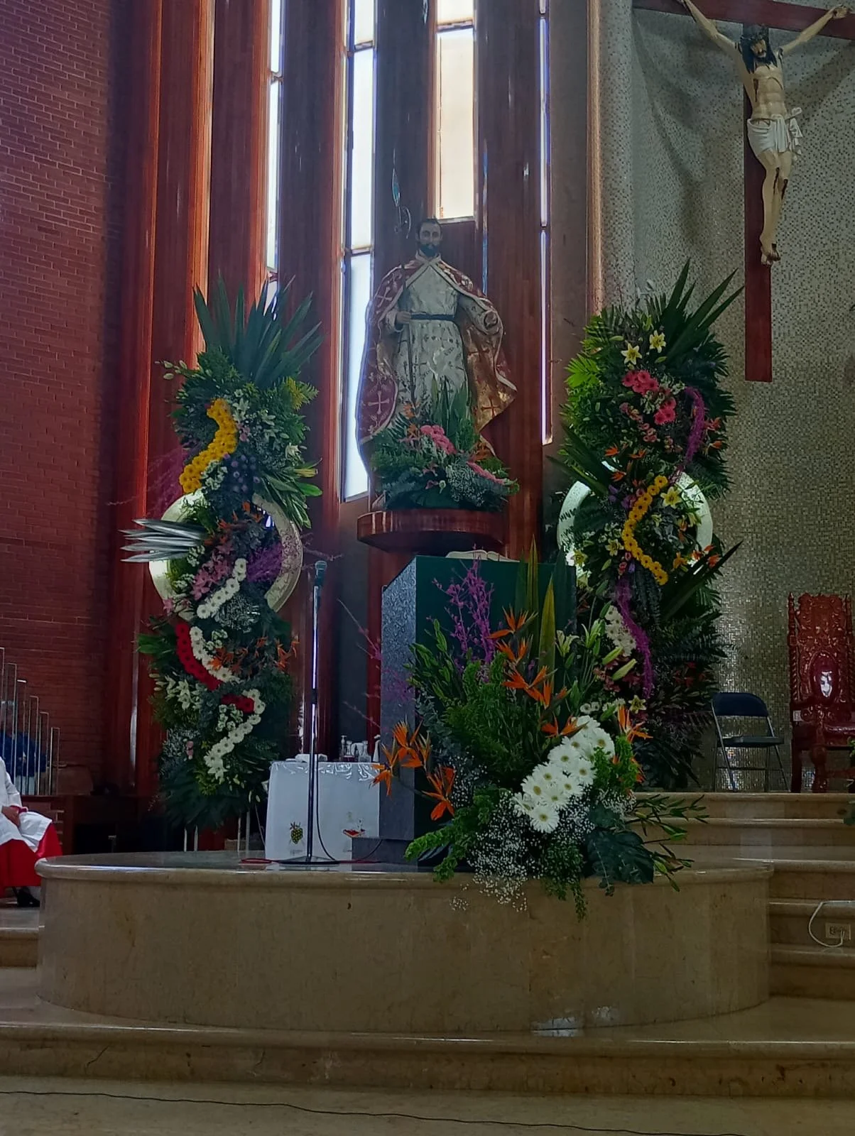 Interior of a church altar decorated with large floral arrangements, a statue of a religious figure at the center, and a crucifix hanging on the wall behind.
