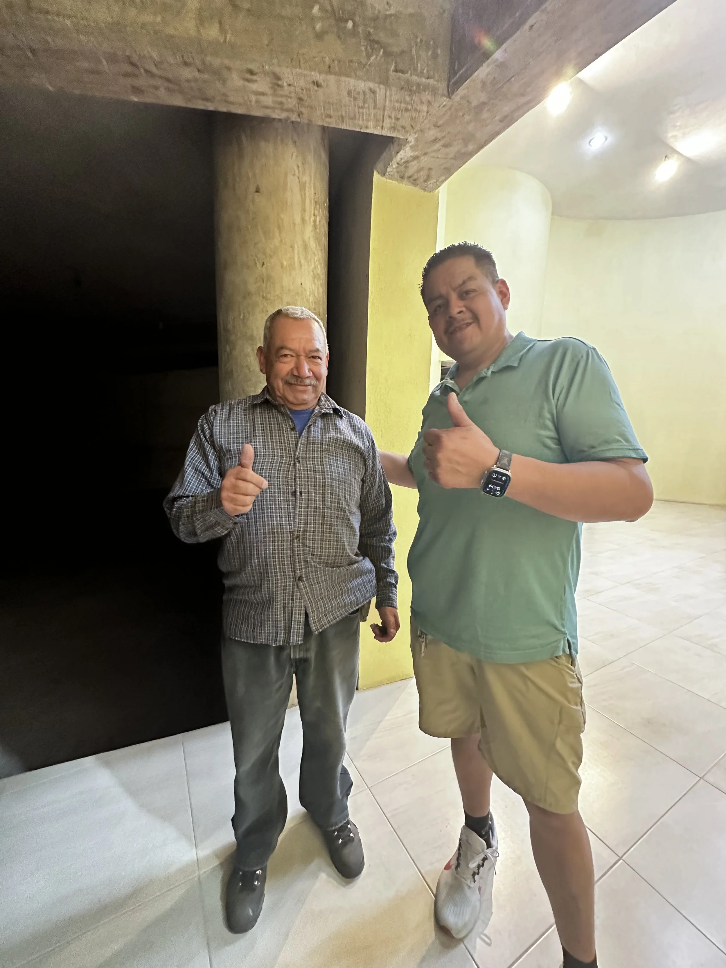 Two men standing indoors, smiling and giving thumbs up, one older with a checkered shirt and the other younger in a mint green polo, beige shorts, and sneakers.