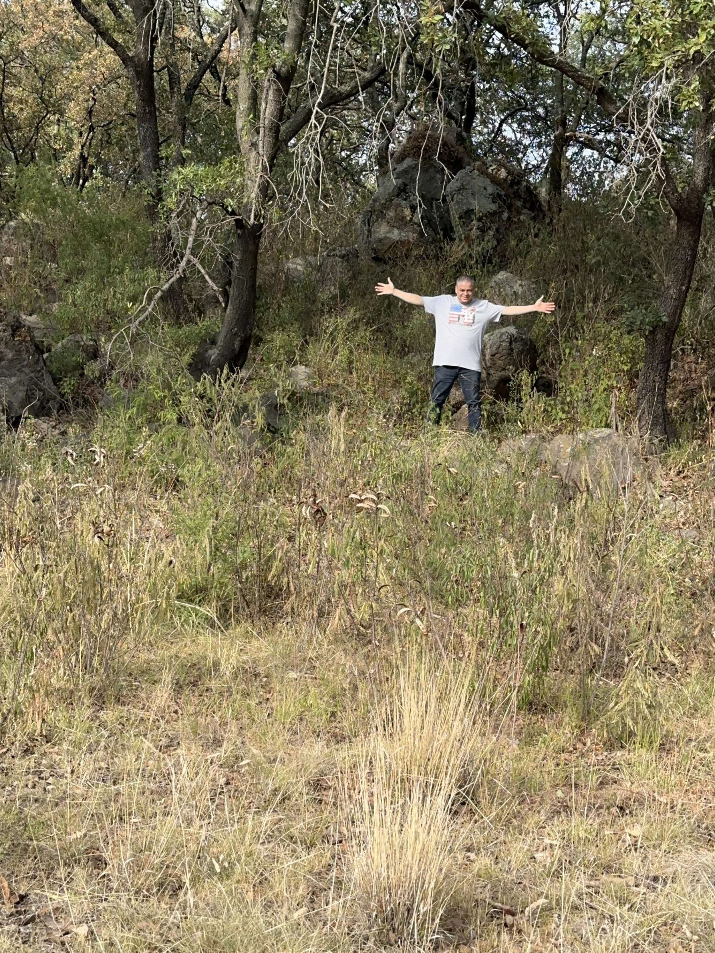 A man standing in a forested area with outstretched arms, surrounded by trees in the pedrerara tlalancaleca tlalankann