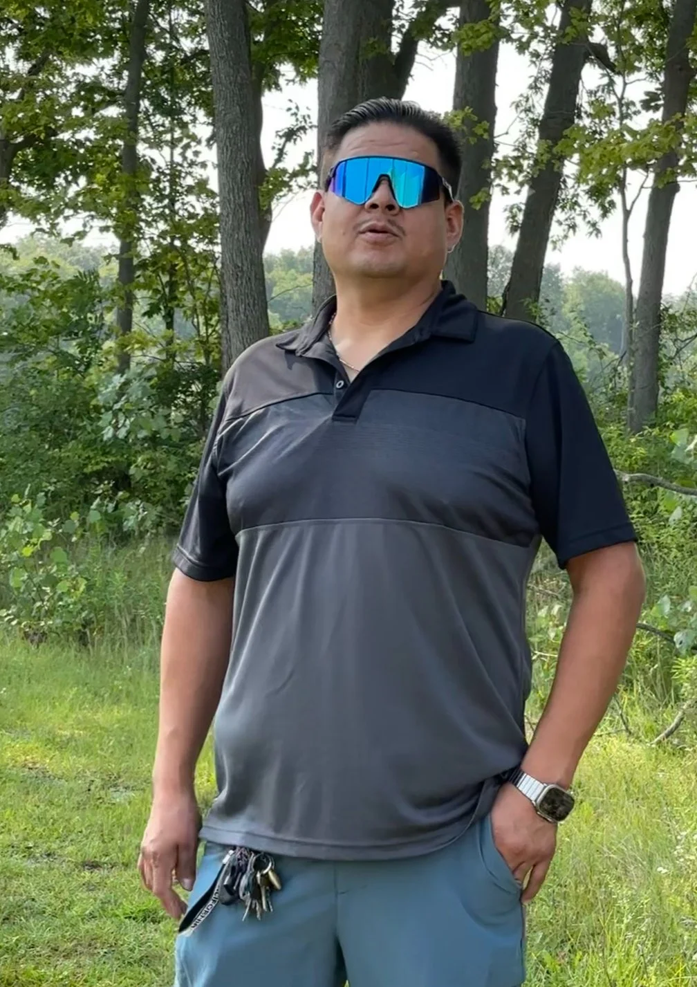 Man in black and gray polo shirt standing outdoors in front of trees, wearing sunglasses and a wristwatch, with a set of keys attached to his shorts.