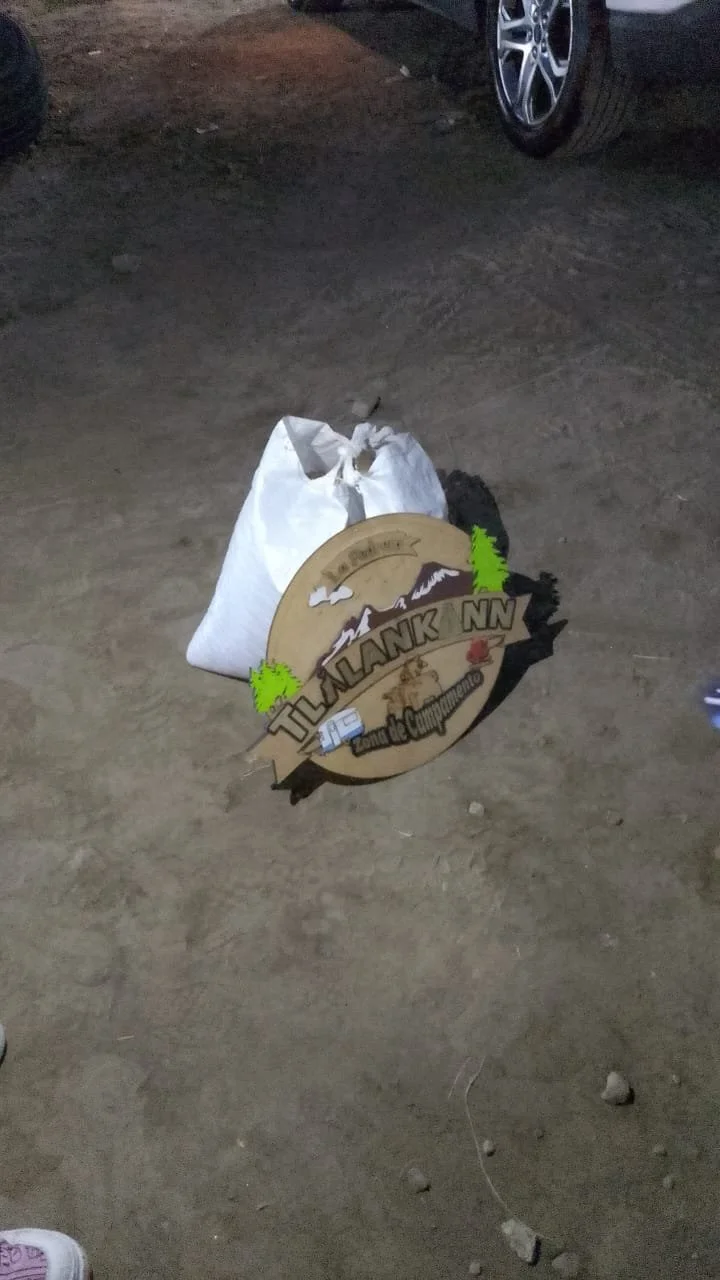 Group of people standing outdoors in a grassy area with trees, some wearing reflective safety vests, and children, with a truck in the background.