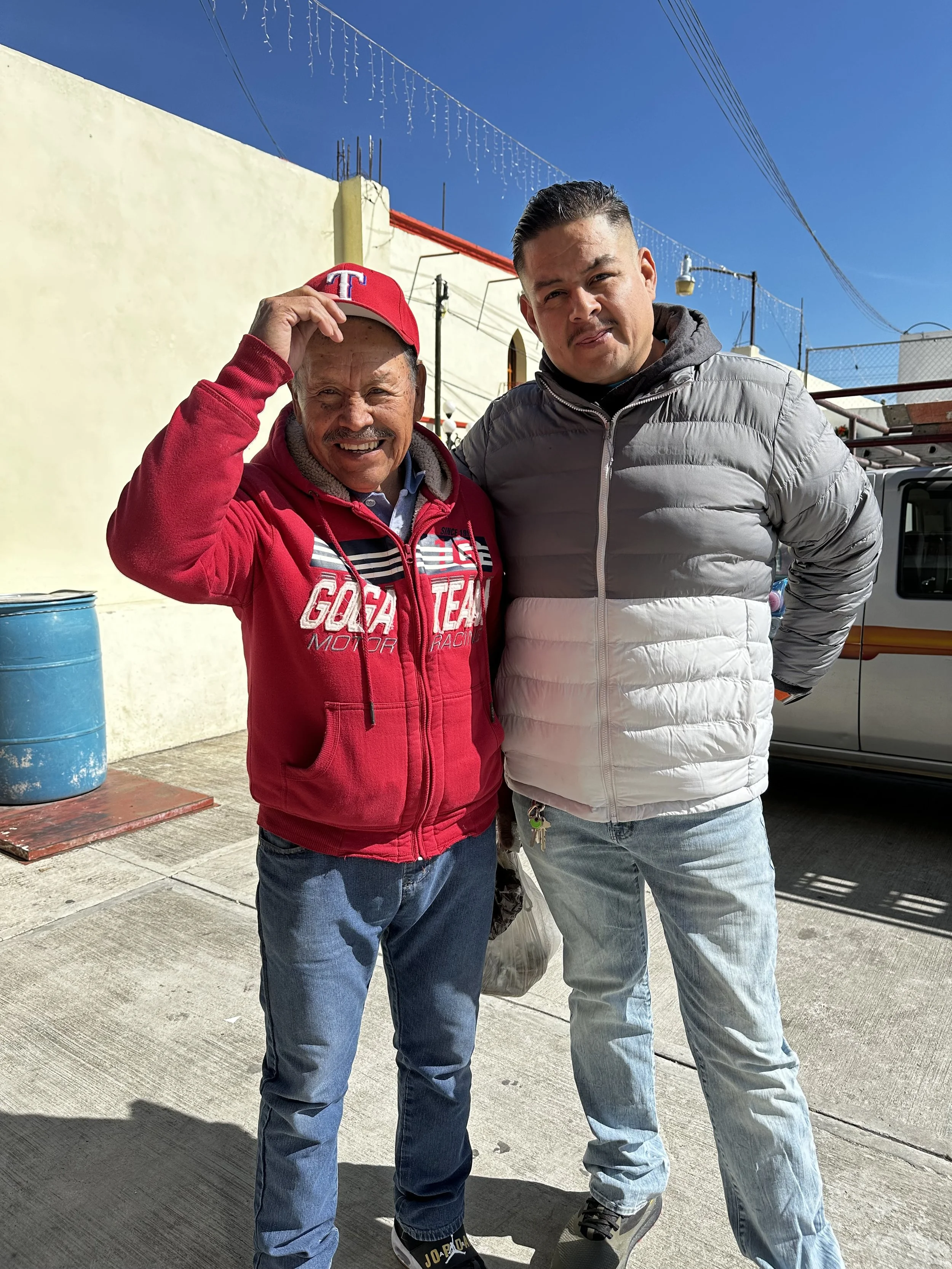 Two men standing outside on a sunny day. The man on the left is wearing a red hoodie, a red cap, and is smiling and holding his cap. The man on the right is wearing a gray puffer jacket and light blue jeans. There are string lights hanging above them, a beige wall, a blue barrel, and a vehicle in the background.