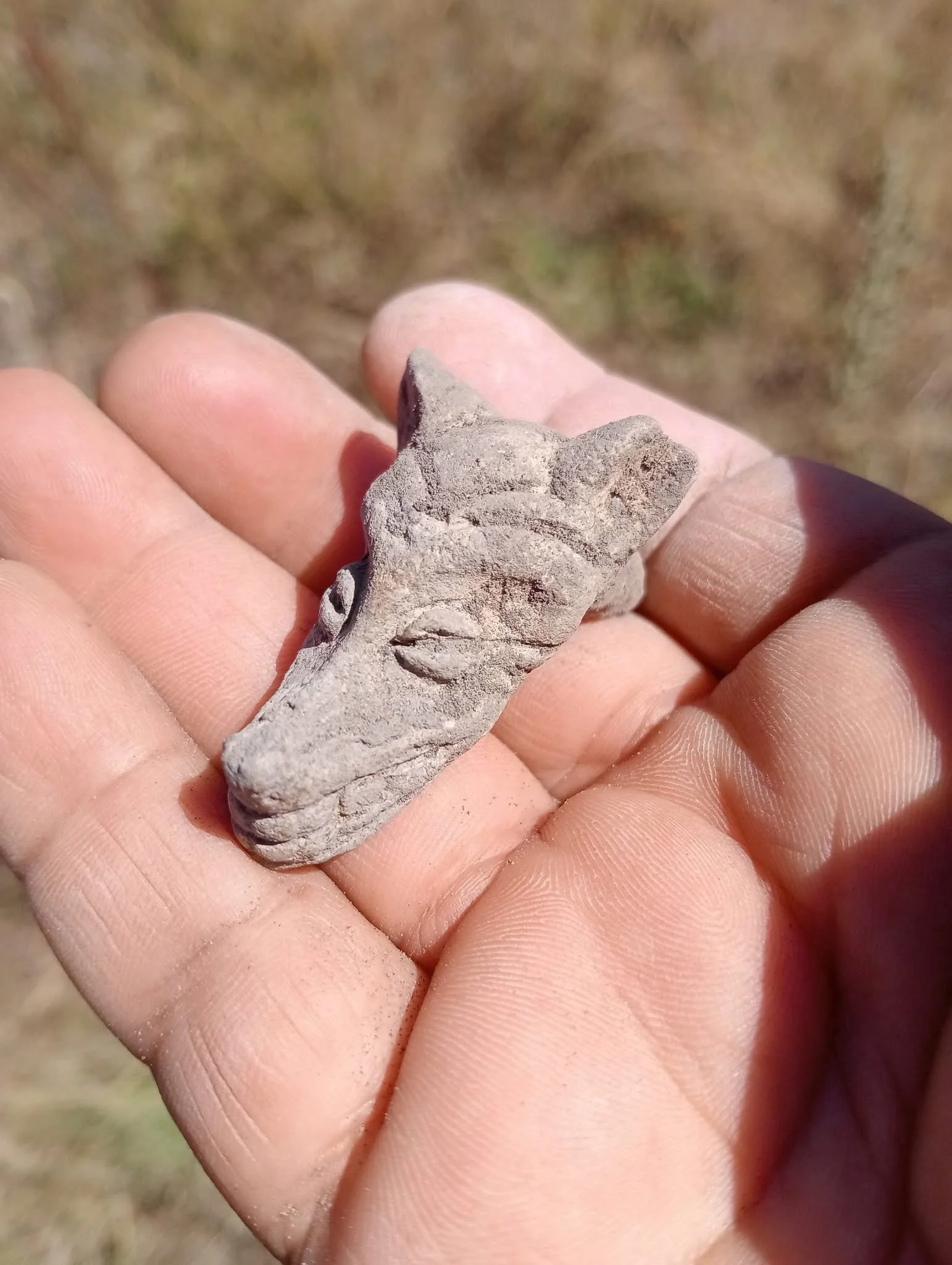 Close-up of a small clay artifact resembling a stylized crocodile head, held in a person's hand outdoors.