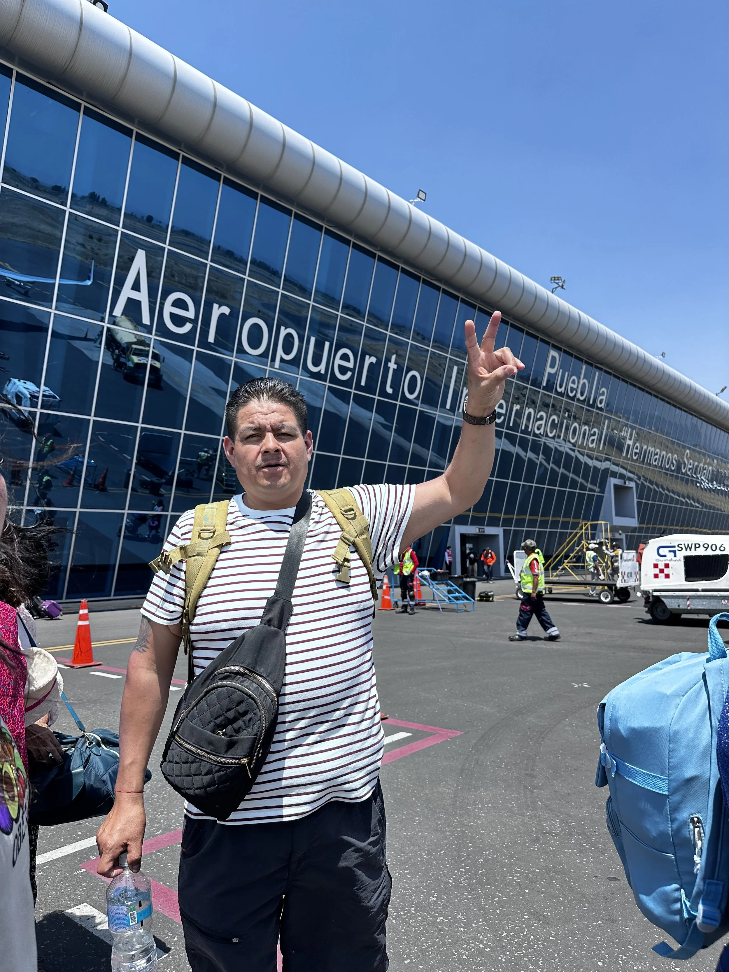 Man with backpack holding water bottle, making a peace sign in front of Aeroport International Hermanos Serdán with glass facade, reflections of airplanes, and airport staff nearby.